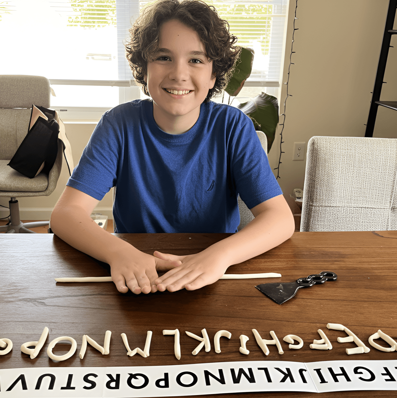 Smiling boy with curly hair sitting at a table with letter tiles, in a bright room with a window and plants.