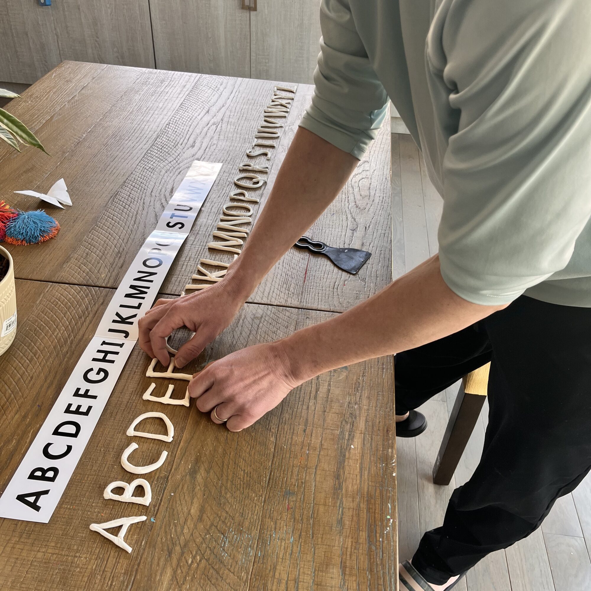 Person arranging a white strip with black letters on a wooden table, with a row of alphabet letters and a small object nearby.