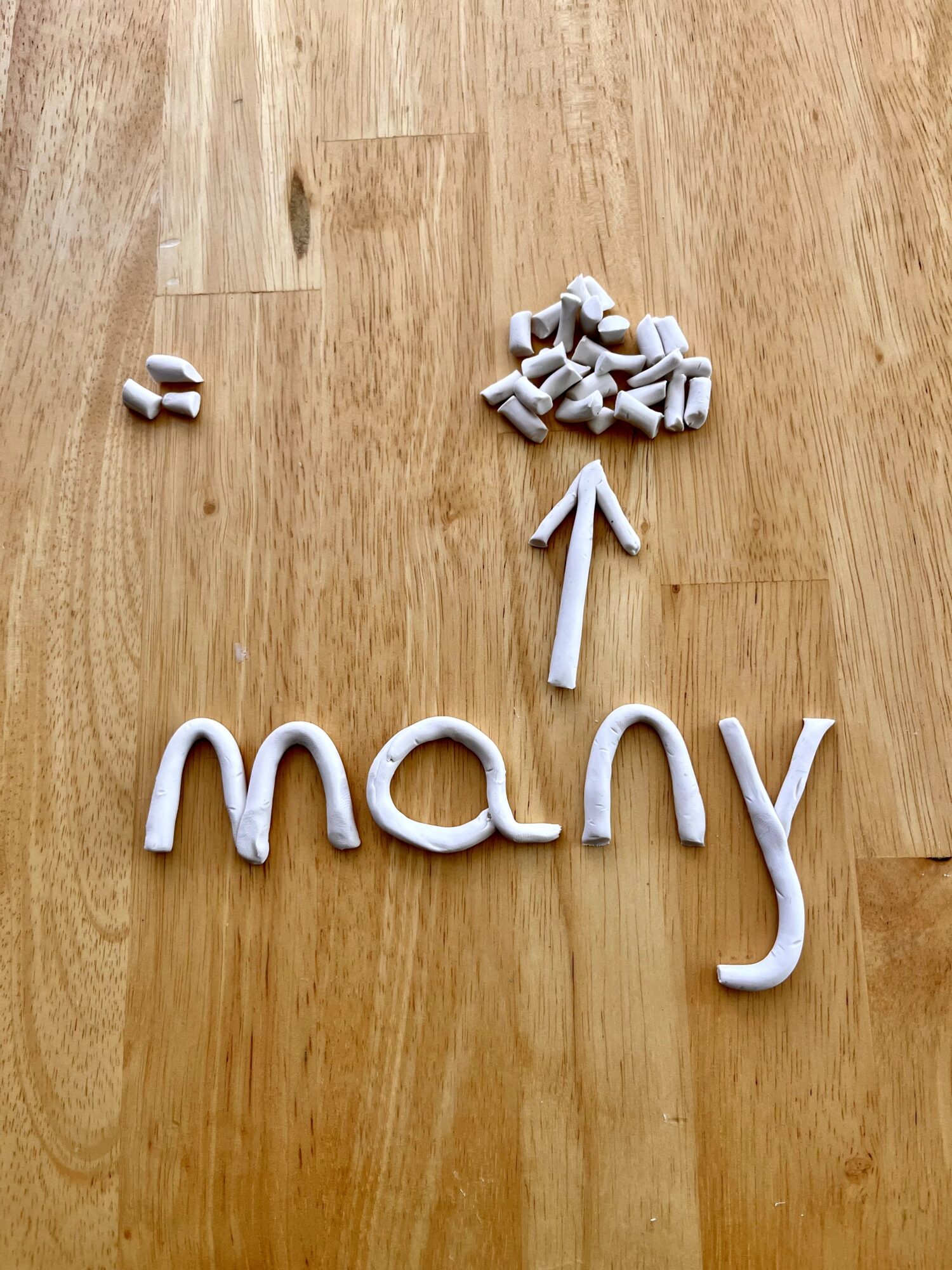 Word 'many' spelled with white clay, with small clay pieces above and an arrow pointing to them, on a wooden surface.