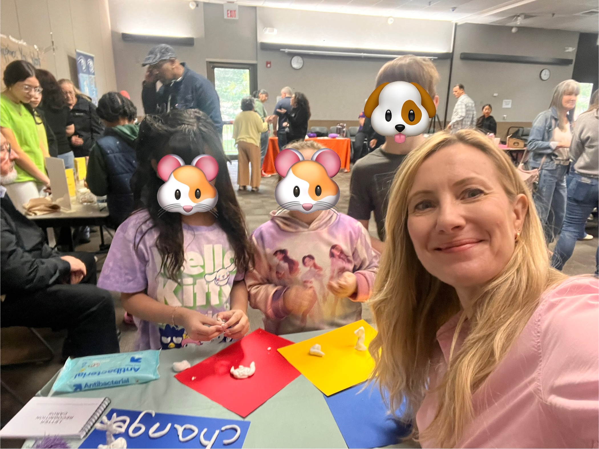 Group of children and adults gathered around a table with colorful paper, in a room with people in the background.