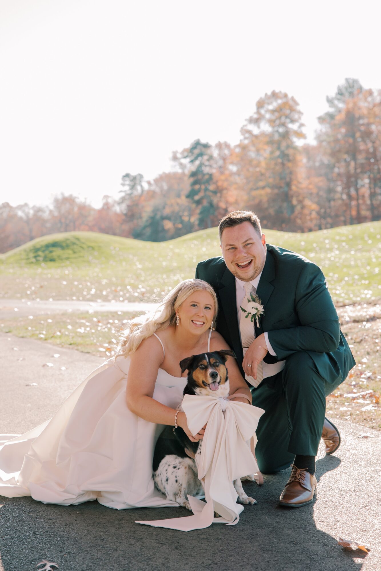 A couple in wedding attire kneels outdoors with a dog, smiling, in a park with trees and grass in the background.