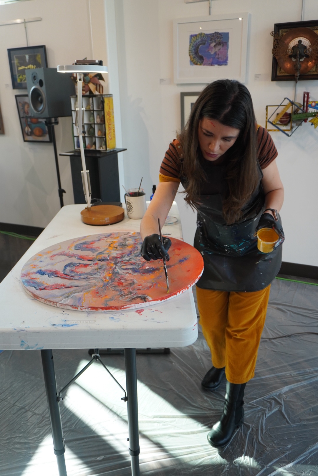 Woman painting on a circular canvas at a table in an art studio, with artwork on the walls behind her.