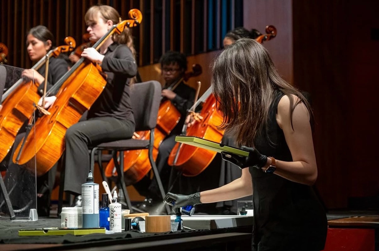 A woman conducts an orchestra of string musicians on stage, with music stands and instruments visible.