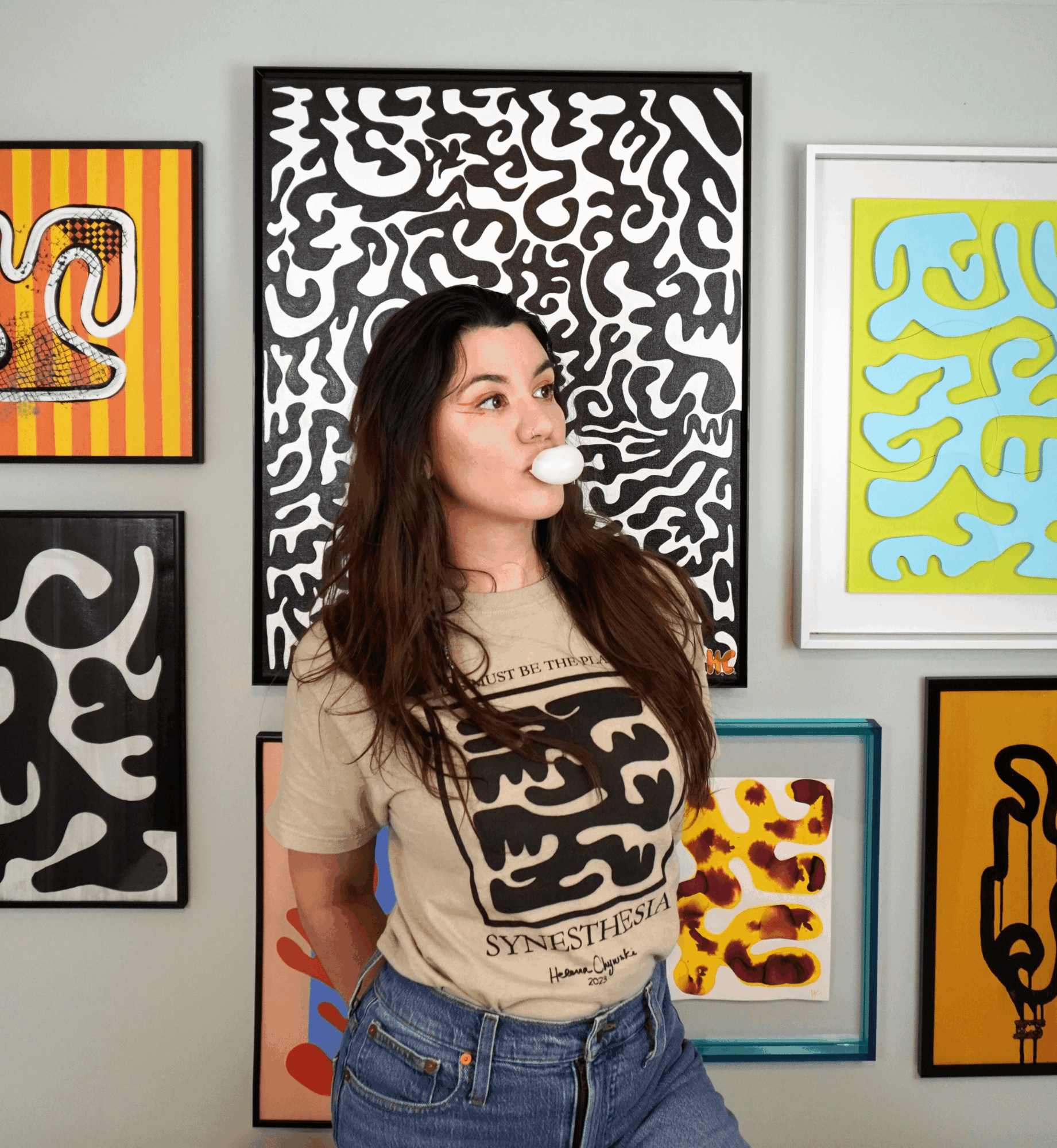 Woman with long dark hair blowing bubble gum, standing in front of abstract black and white artwork, wearing a beige T-shirt and jeans.