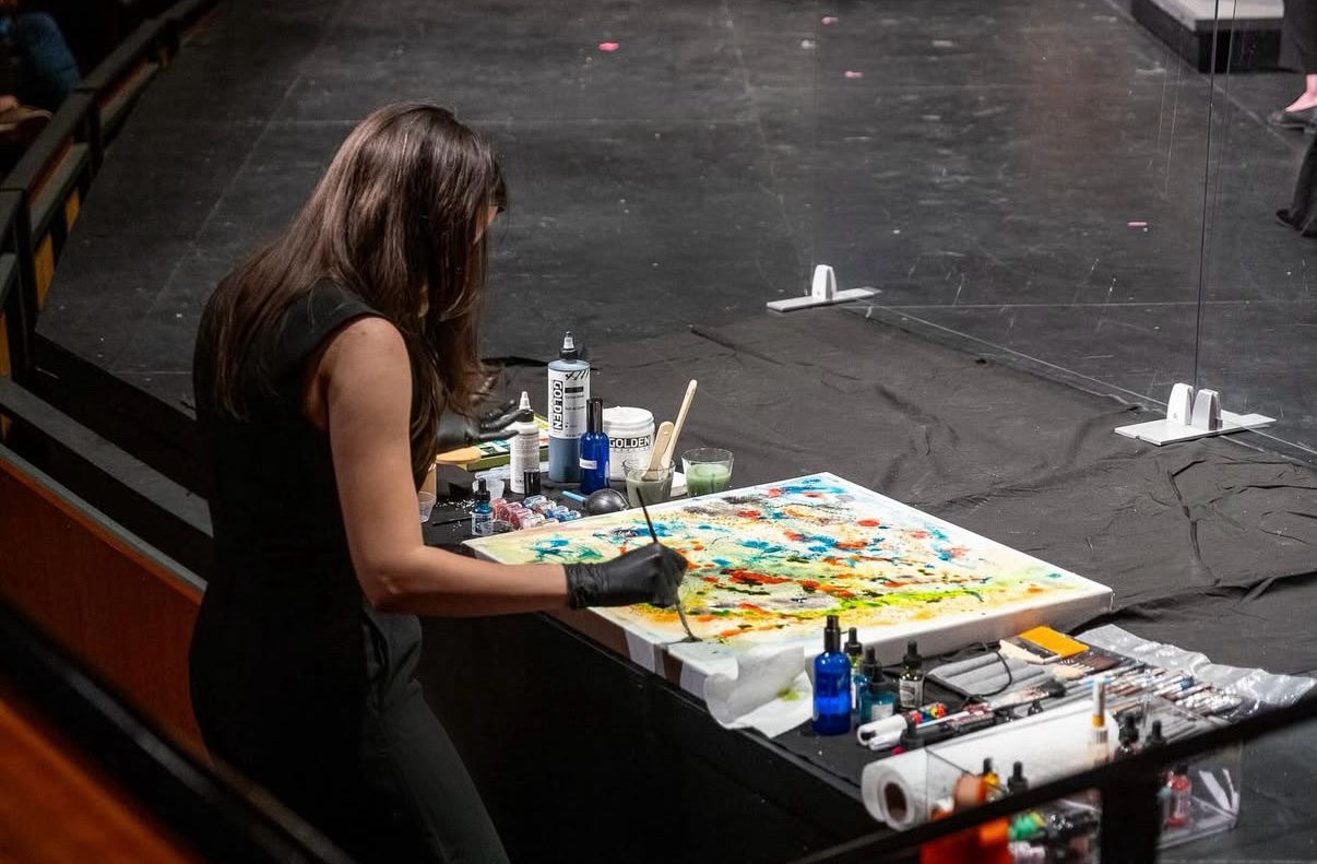 Woman painting on a large canvas in a dark room, surrounded by art supplies and bottles.