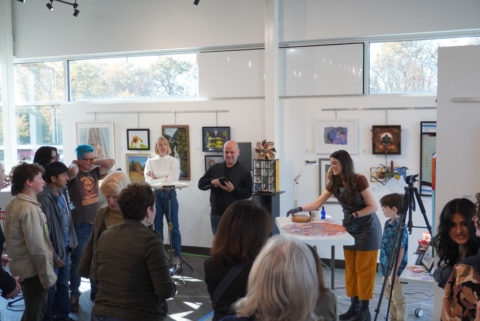 Group of people gathered indoors, some listening to a person speaking, with artwork displayed on white walls and large windows.
