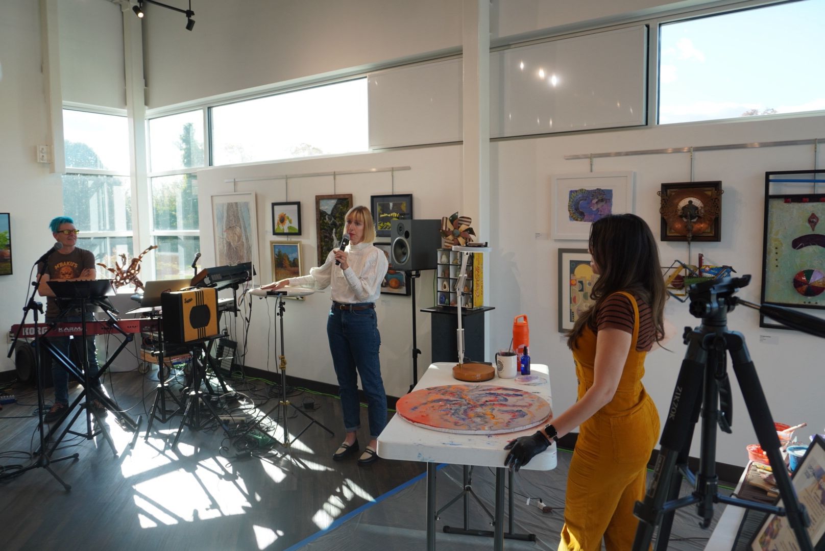People in an art gallery with paintings on the wall, a woman speaking, and a woman at a table with art supplies.