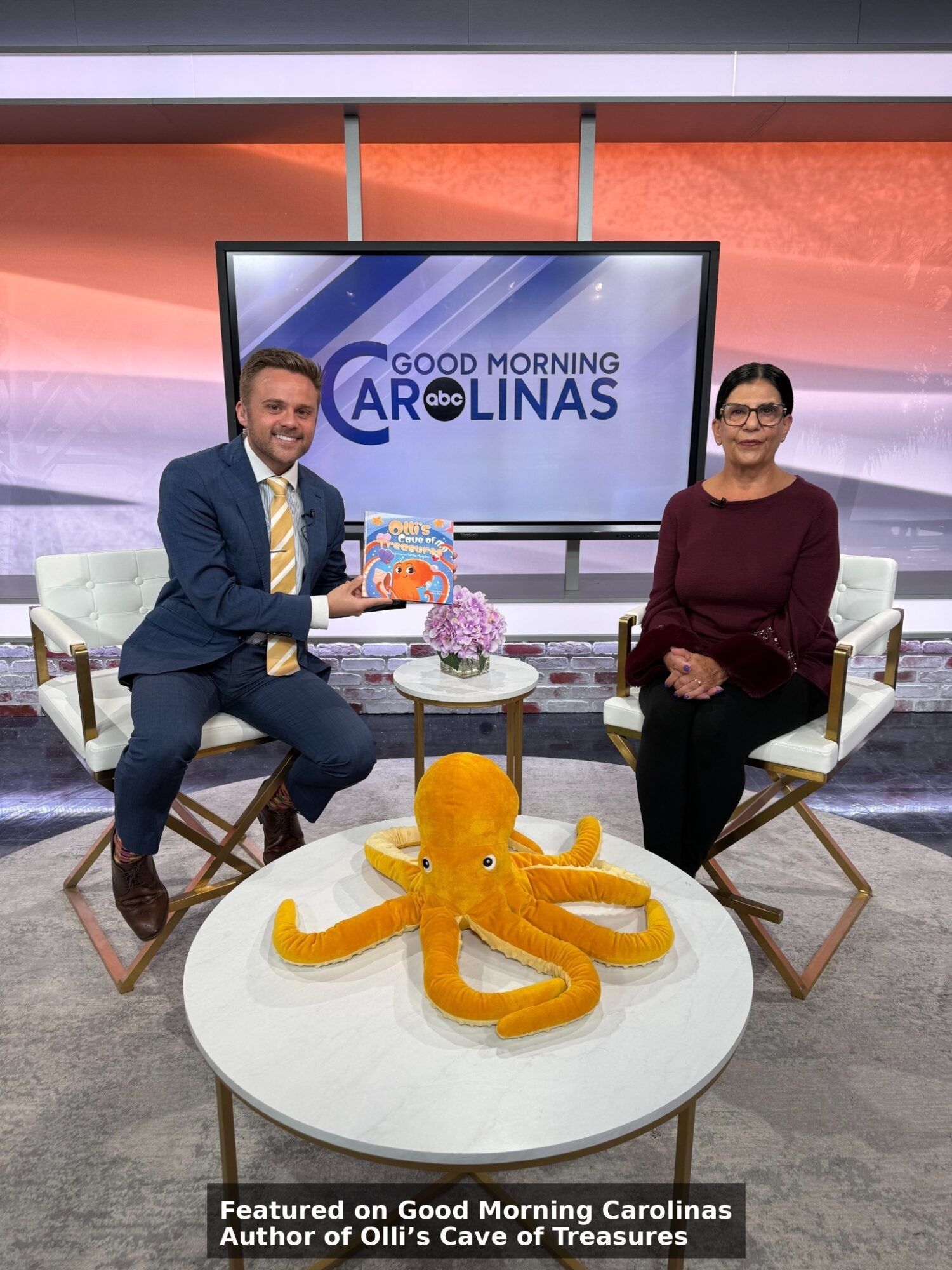 Two people sit on chairs in a TV studio with a large screen behind them displaying 'Good Morning Carolinas.' A plush orange octopus toy is on a table in front of them.
