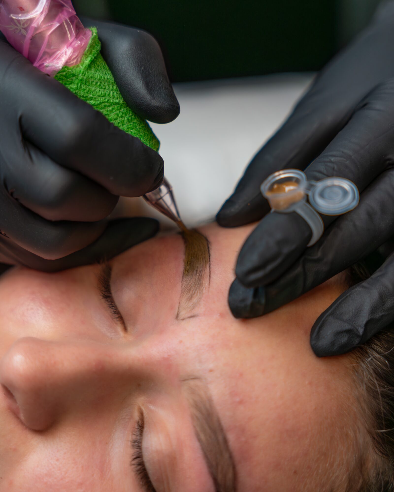 Person receiving eyebrow tattooing, technician wearing black gloves, holding tattoo equipment near closed eye, skin prepared for procedure.