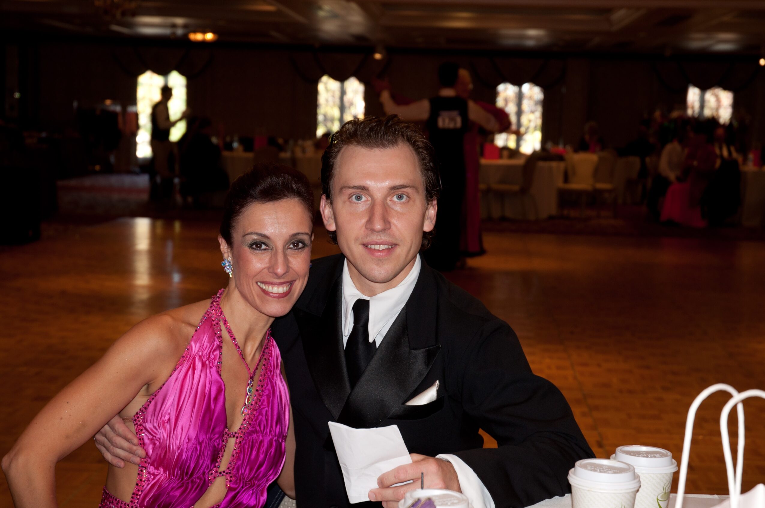 A woman in a pink dress and a man in a black suit sit together at a formal event, smiling at the camera.