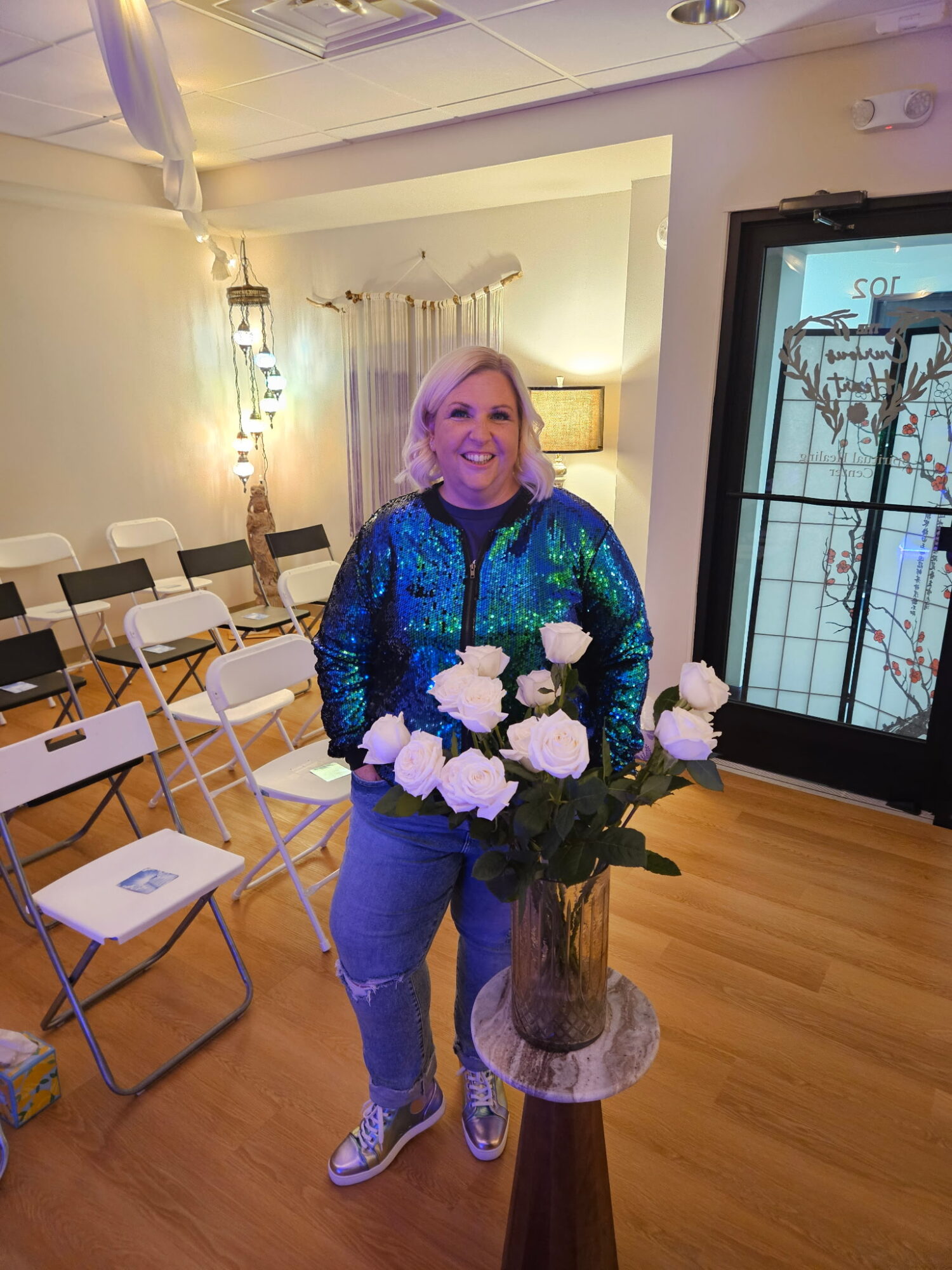 Woman with blonde hair smiling, standing behind a bouquet of white roses in a vase, in a room with chairs and a door with glass panes.