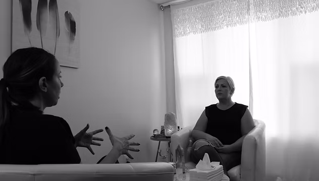Two women sitting and talking in a room with a window and curtains, one gesturing with hands.