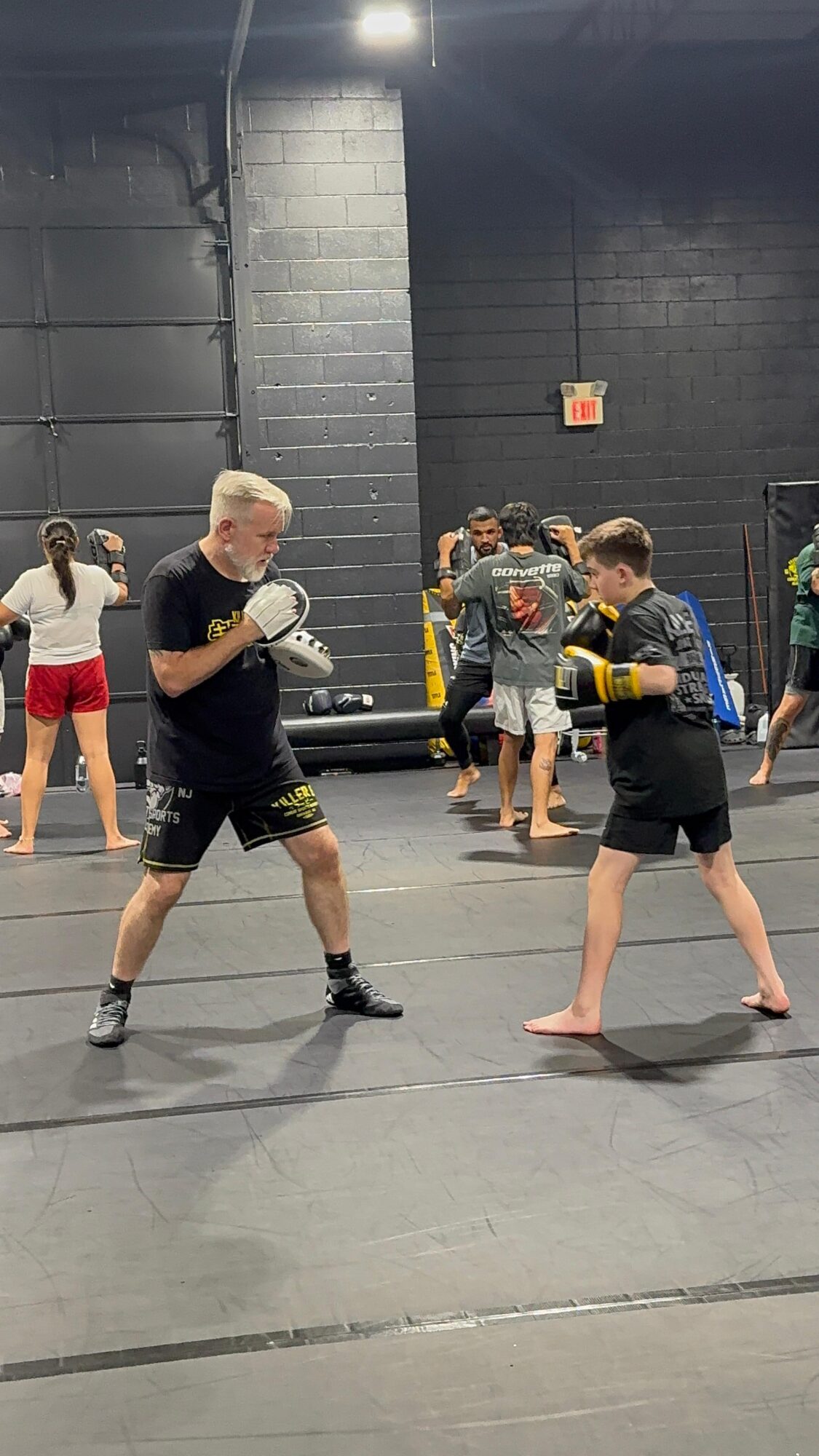 Two people practicing boxing in a gym, one with gloves and pads, others in the background, some watching or preparing.