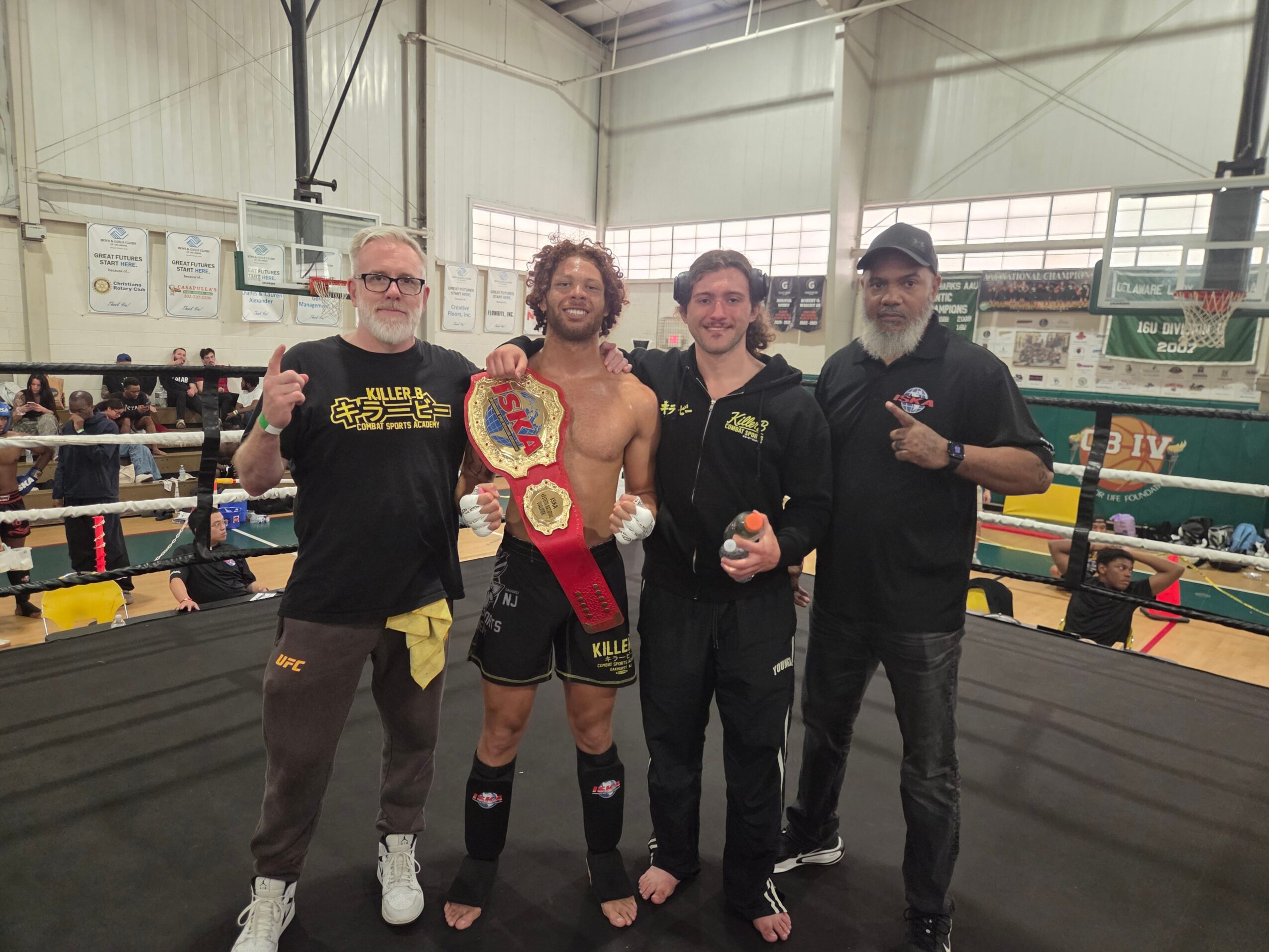 Four men standing in a boxing ring, one shirtless with a championship belt, others in casual clothing, inside a gym.