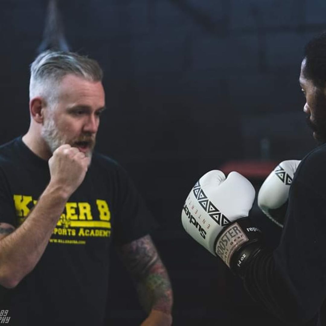Two men in a boxing gym, one with boxing gloves, face each other during training, dark background, focused expressions.