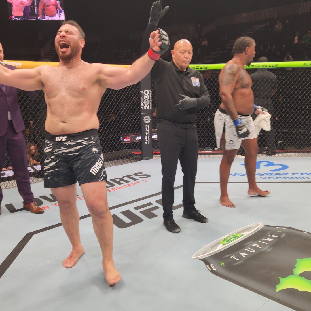 Celebrating mixed martial arts fighter with arms raised, referee and opponent in the background inside the octagon.
