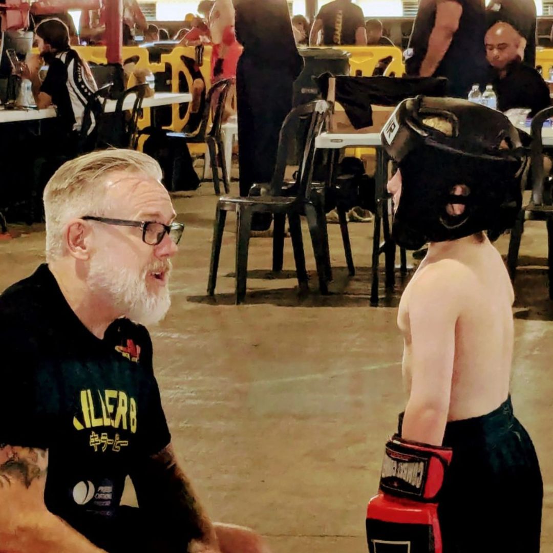 A shirtless child wearing a black helmet and red boxing gloves faces an older man with glasses and a beard, indoors.