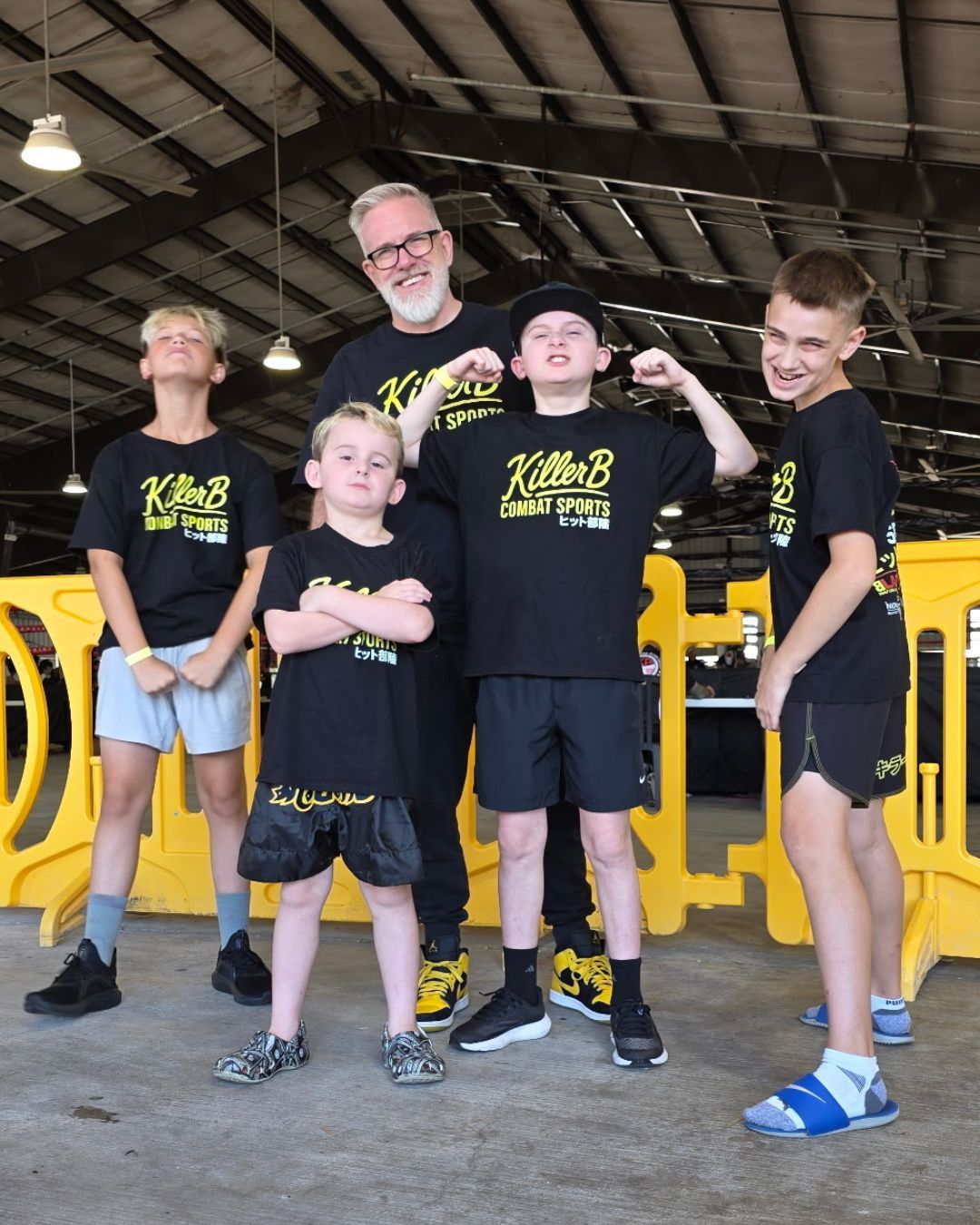 Group of five people, including an adult man and four children, standing inside a building with a yellow vehicle in the background.
