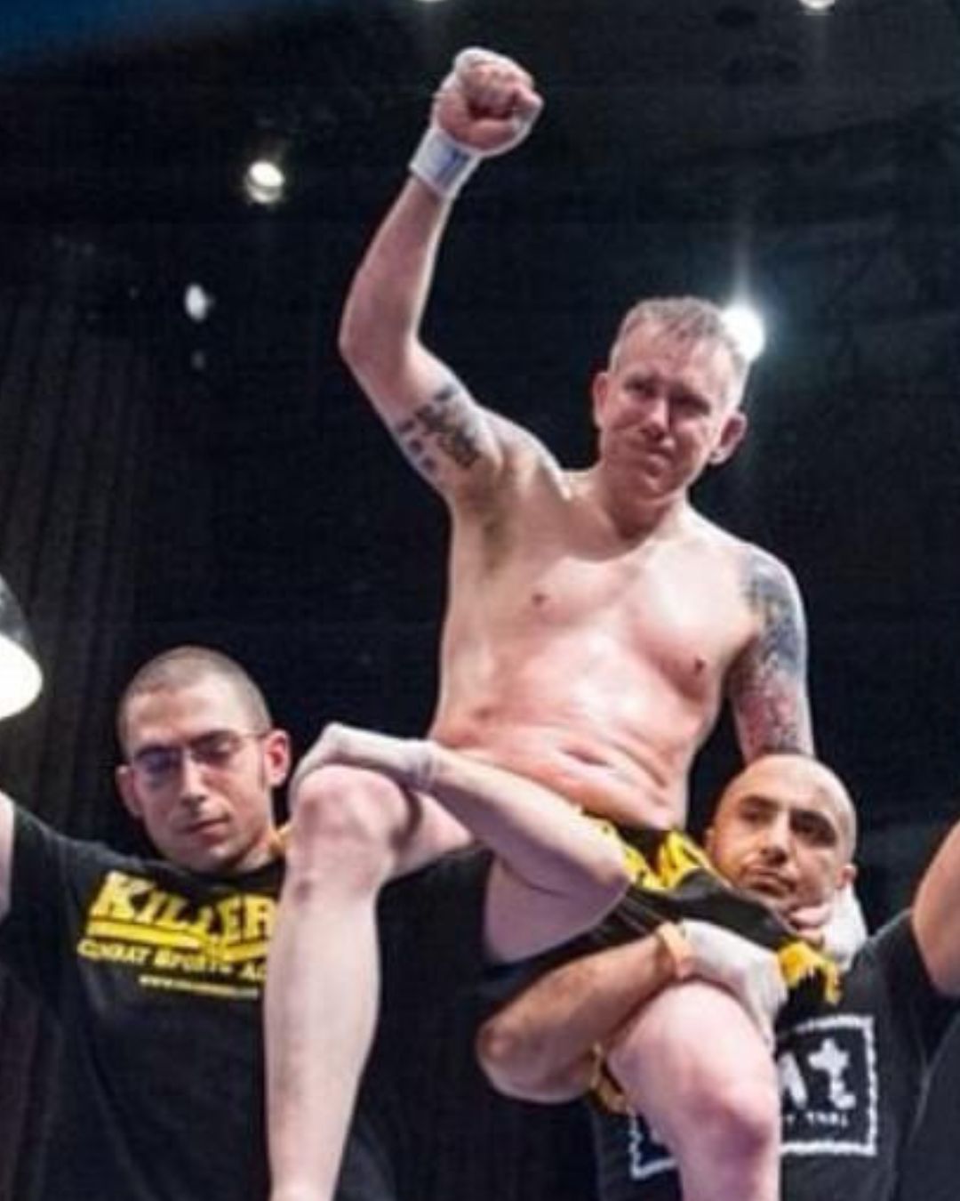 A shirtless man with tattoos raises his fist in the air, supported by two others wearing black shirts with yellow text.