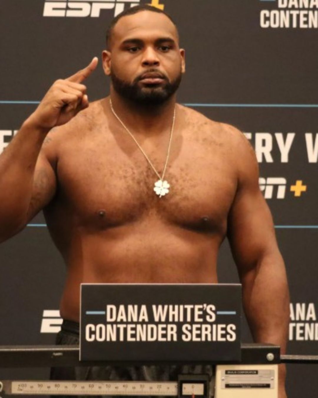 Man with a beard and muscular build pointing to his head, standing behind a sign that reads Dana White's Contender Series.
