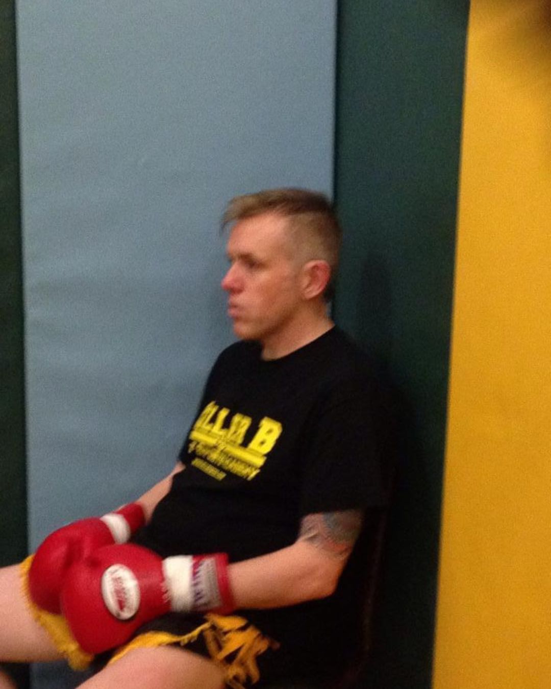 Person wearing boxing gloves sitting against a wall, with a focused expression, in a gym setting.