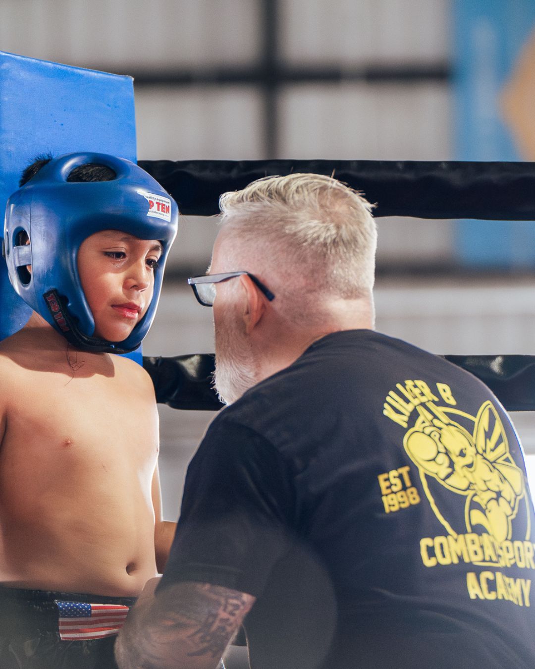 A young boy wearing a blue boxing helmet listens to a coach inside a boxing ring. The coach has short blonde hair and glasses.