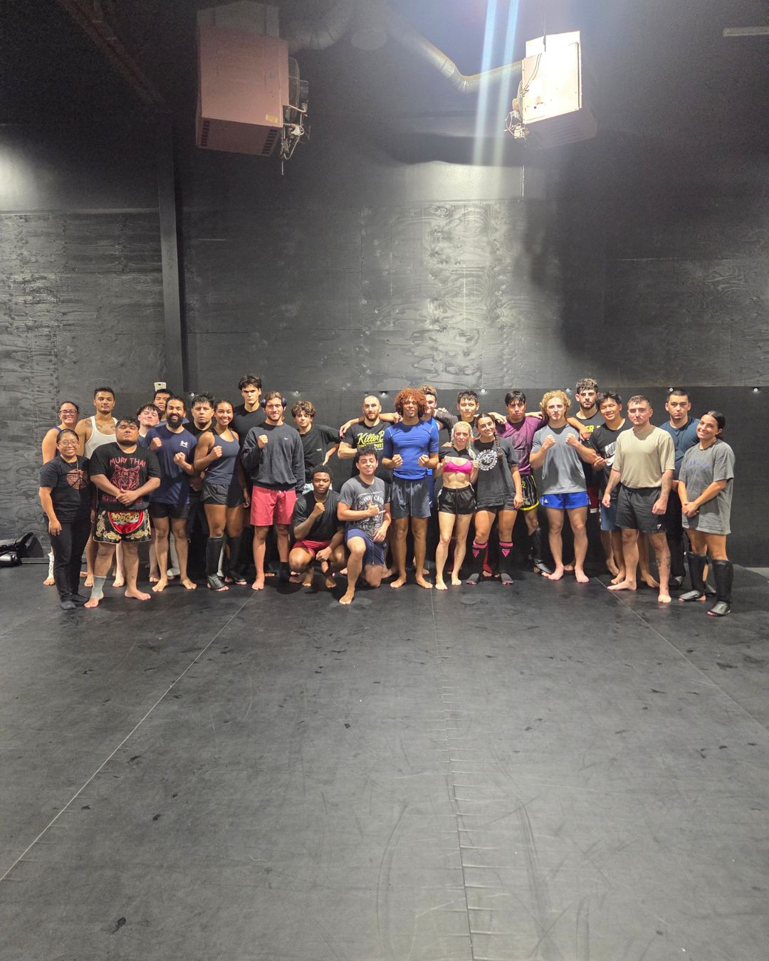 Group of people standing together in a dark indoor space, some in athletic clothing, posing for a photo.