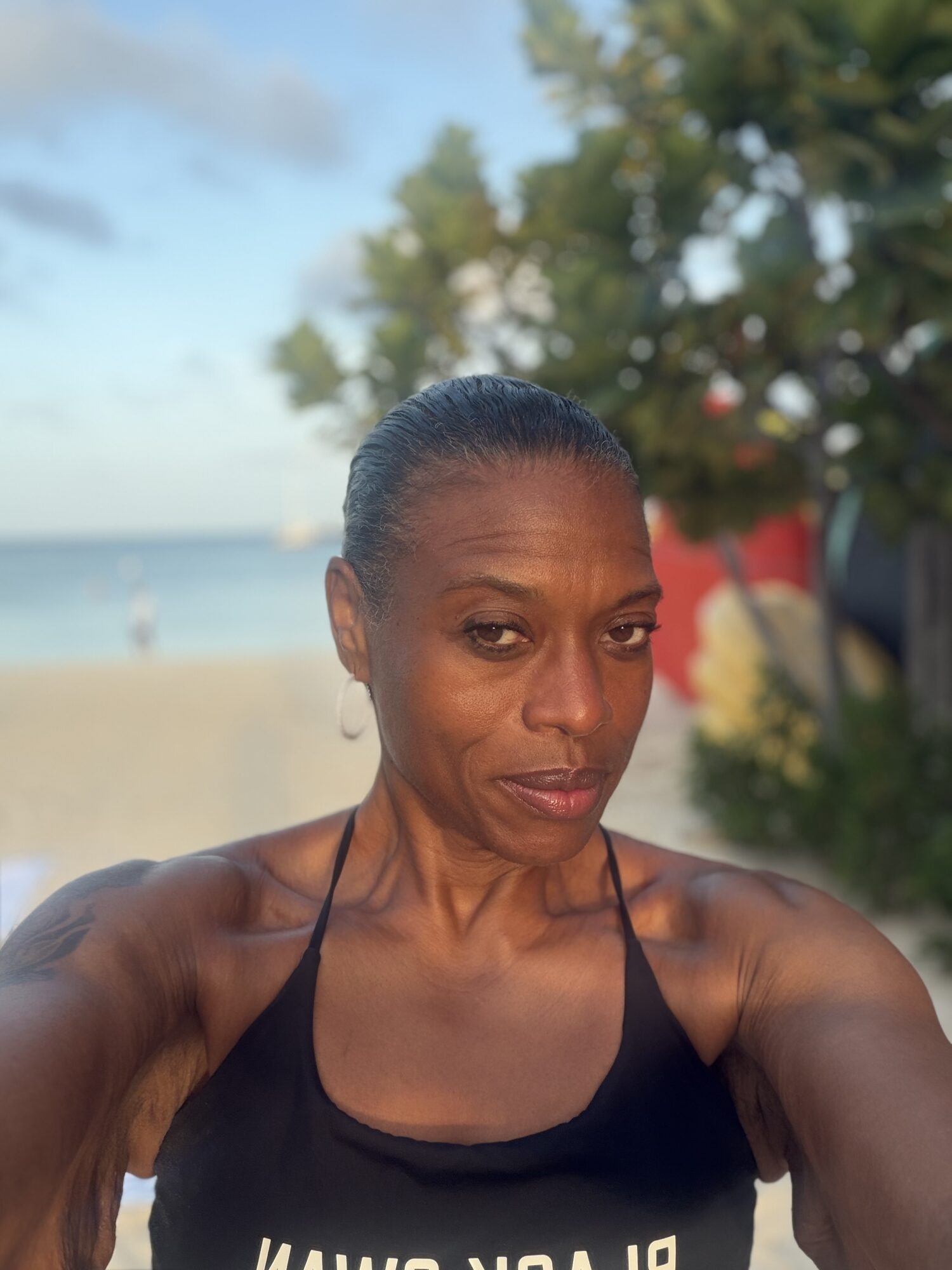 Woman taking a selfie outdoors near water with trees and sky in background, wearing a black top, smiling slightly.