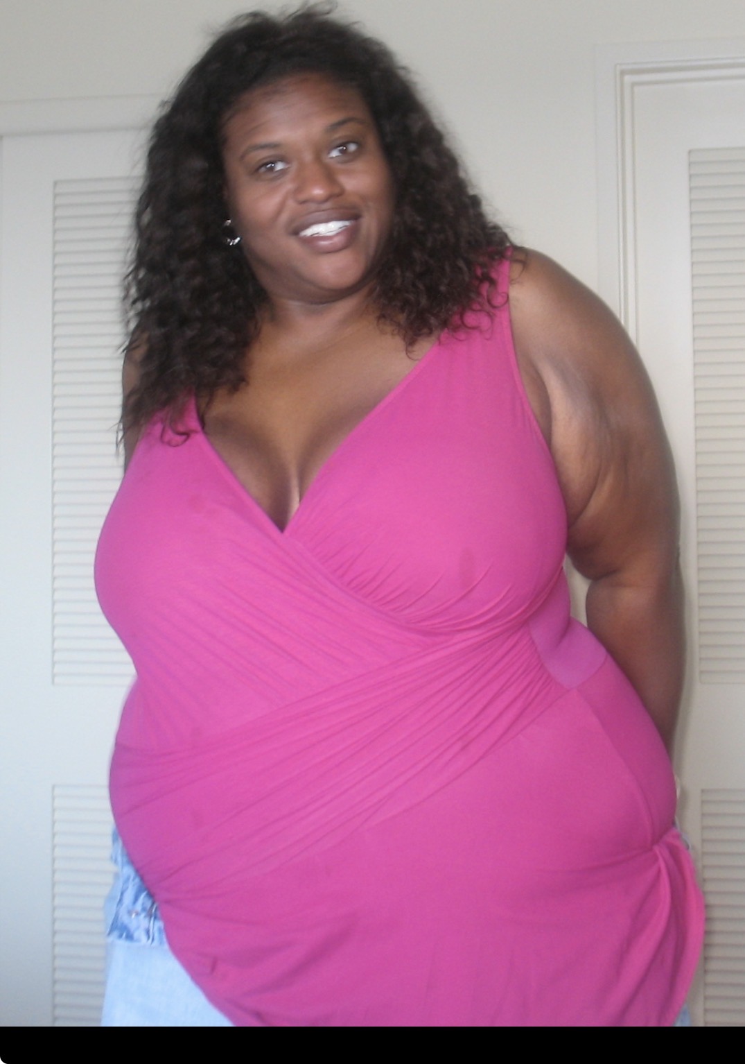 Woman with curly hair wearing a pink sleeveless top, smiling, standing indoors with a white door in the background.