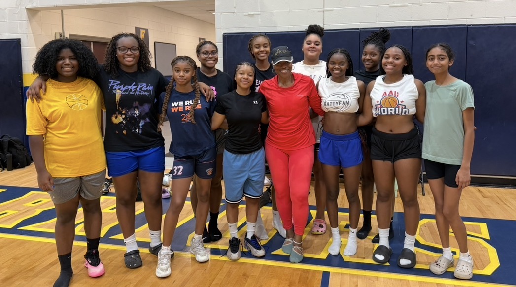 Group of ten young women standing together in a gymnasium, smiling, with a basketball court floor visible.