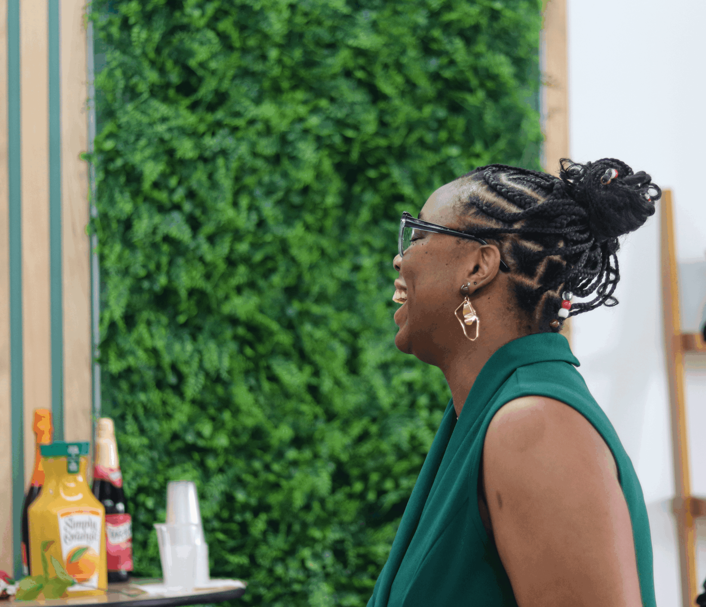 Side view of a smiling woman with braided hair and glasses, wearing a green sleeveless top, standing indoors near a green wall.