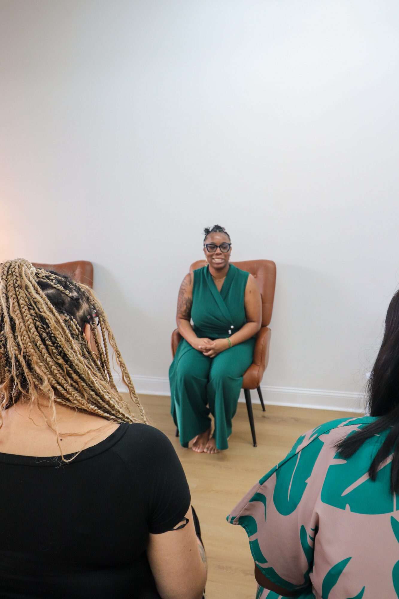 Woman sitting on a chair, wearing glasses and a green outfit, in a room with plain white walls.