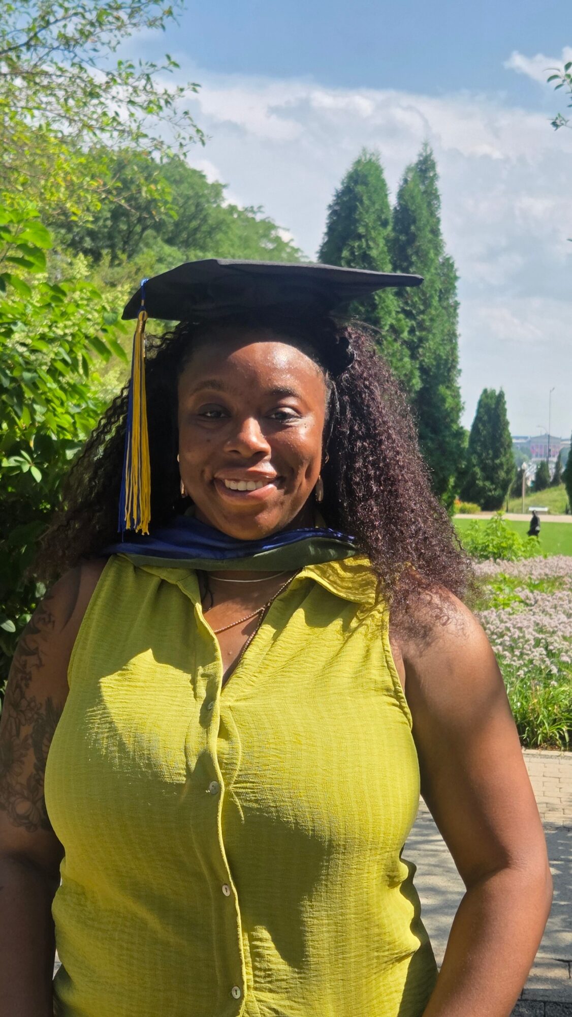 Woman in graduation cap outdoors, smiling, wearing a yellow sleeveless top, greenery and trees in background.
