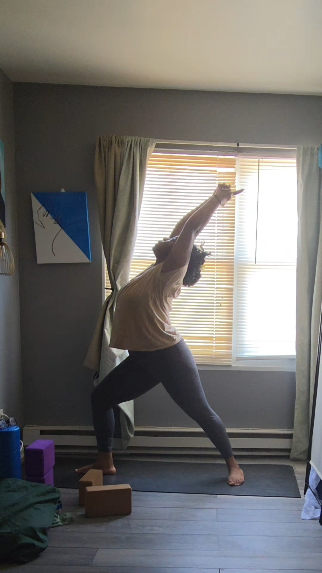 Person practicing yoga in a room with large window, curtains, and wooden floor, stretching arm upward, facing window.