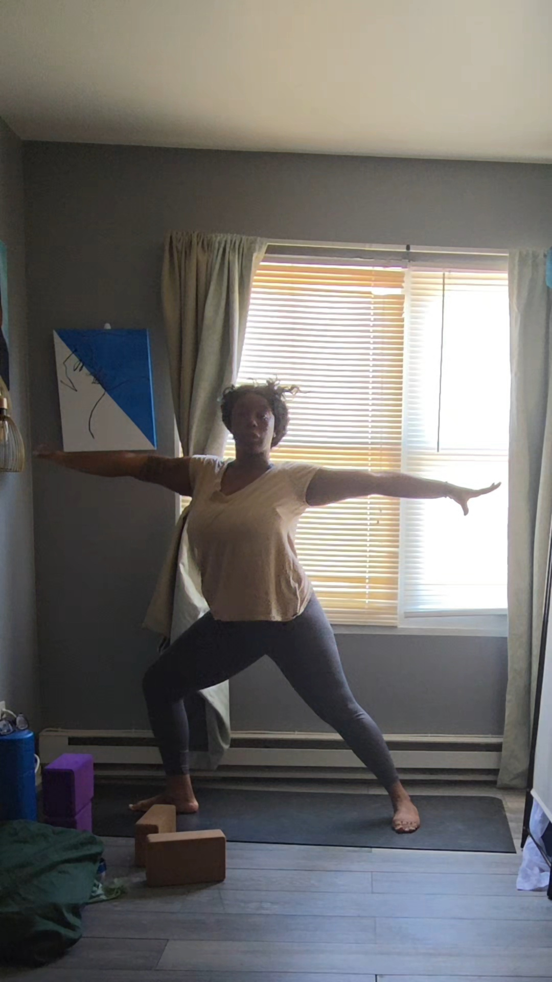 Person standing in a yoga pose with arms extended, in front of a window with blinds and curtains, in a room.