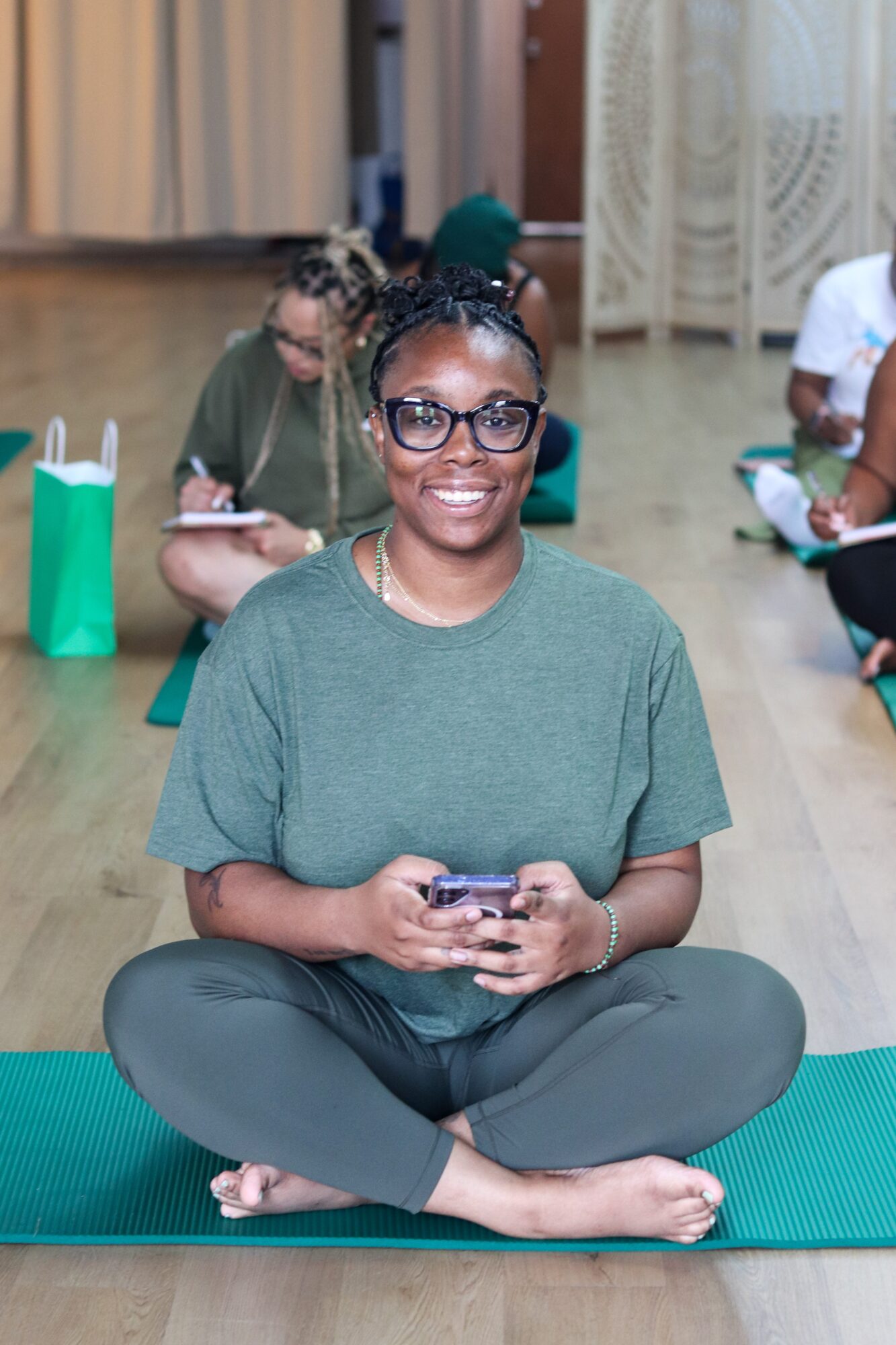 Smiling woman sitting cross-legged on a yoga mat, holding a phone, in a room with other people and curtains.