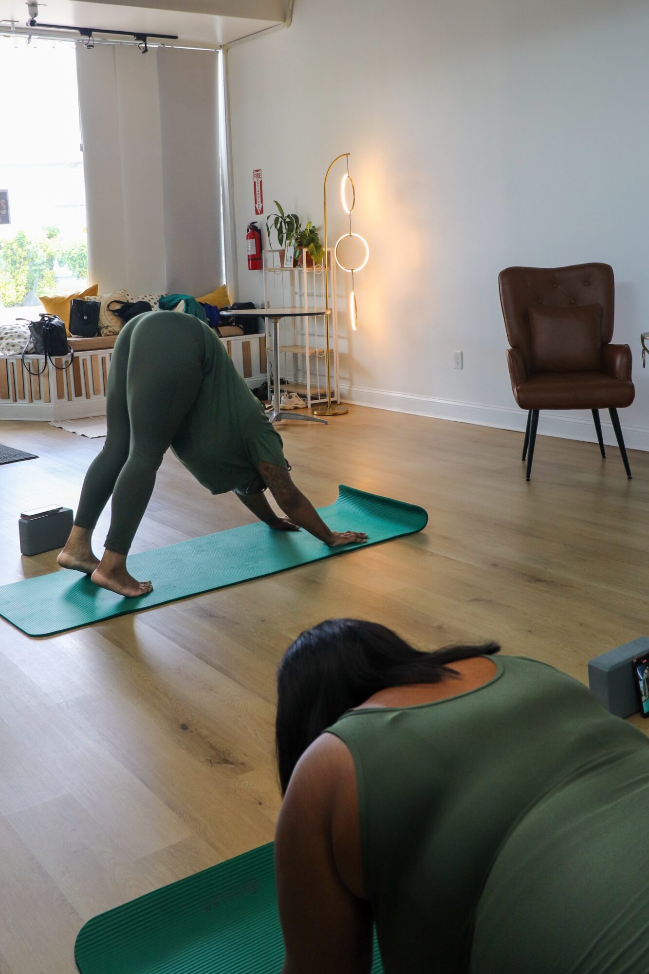 Person practicing yoga on a green mat in a bright room with a window, chair, and decorative lamp, facing downward with hands and feet on the mat.