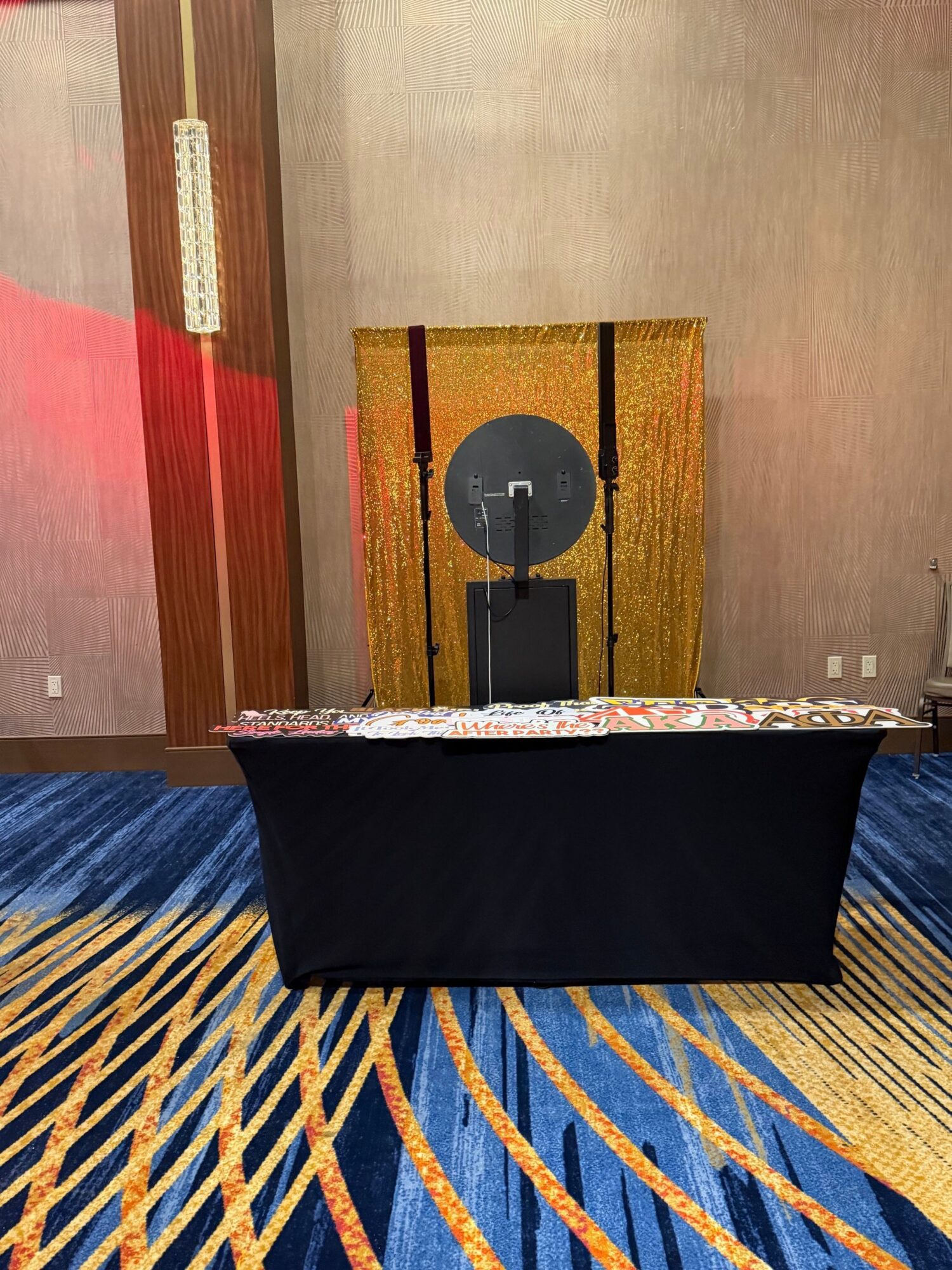 Table with a black tablecloth, a floral table runner, and a black speaker on a wall with gold and red curtains, and a patterned carpet.