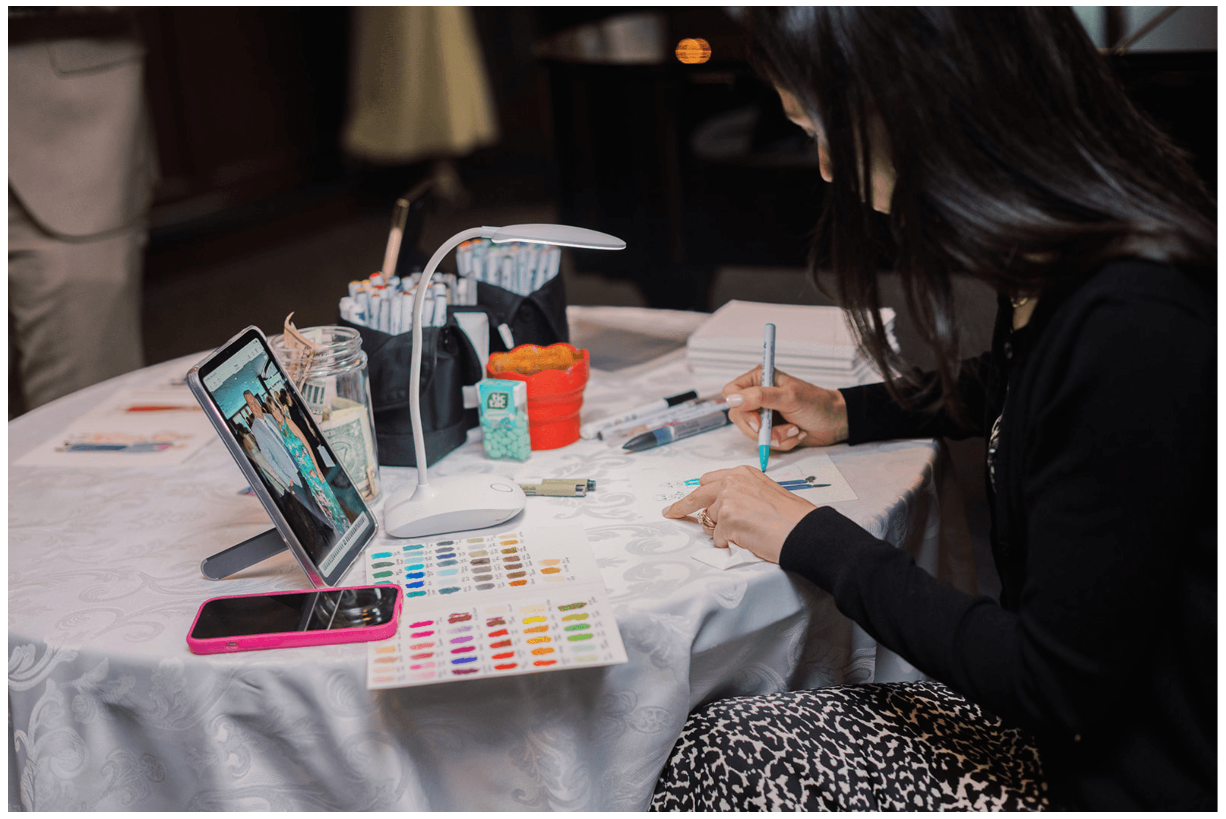 Person painting nails at a table with nail polish bottles, color palette, and a tablet, with long dark hair and black clothing.
