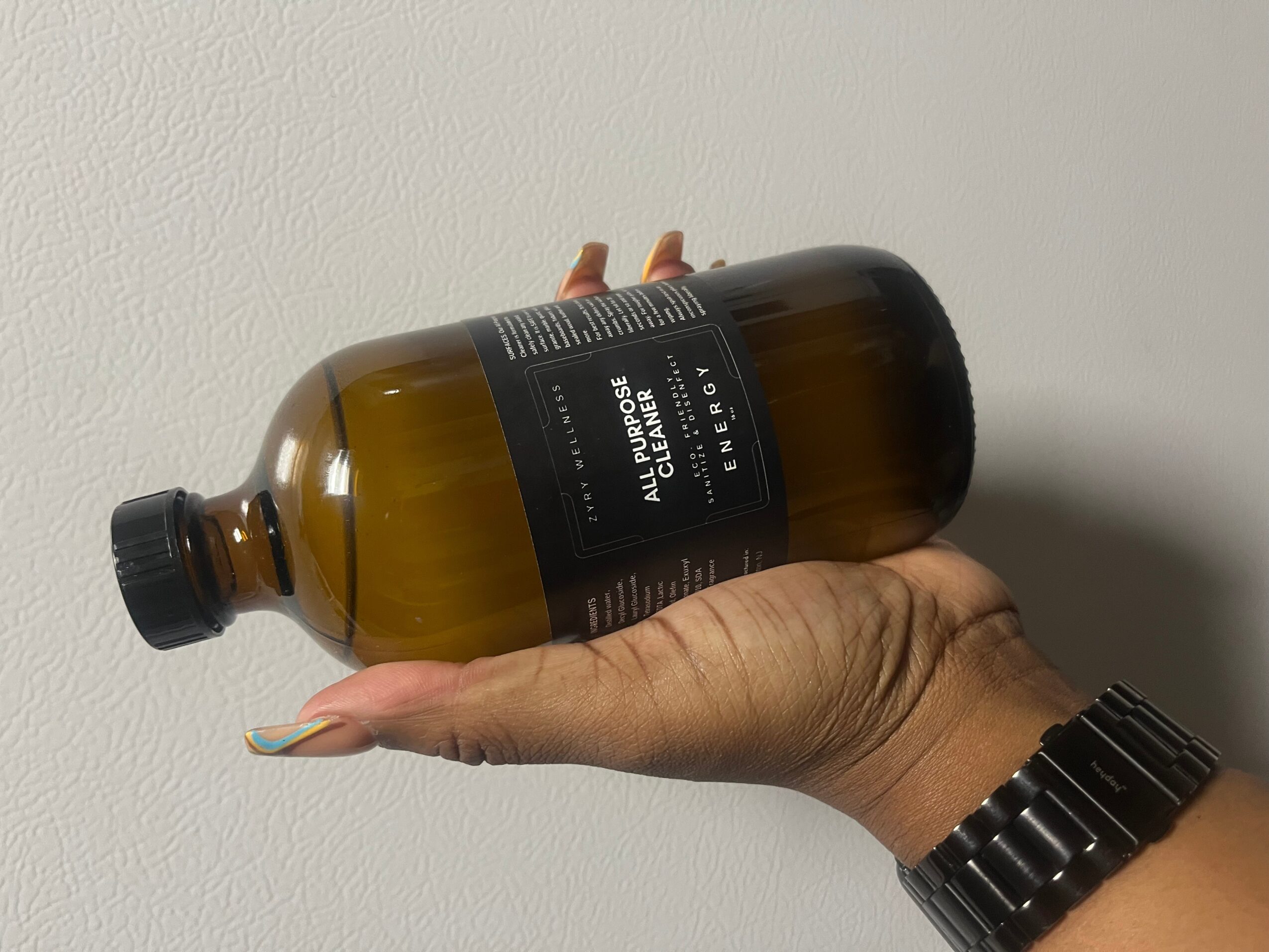 Hand holding a large amber glass bottle with a black label against a plain wall.