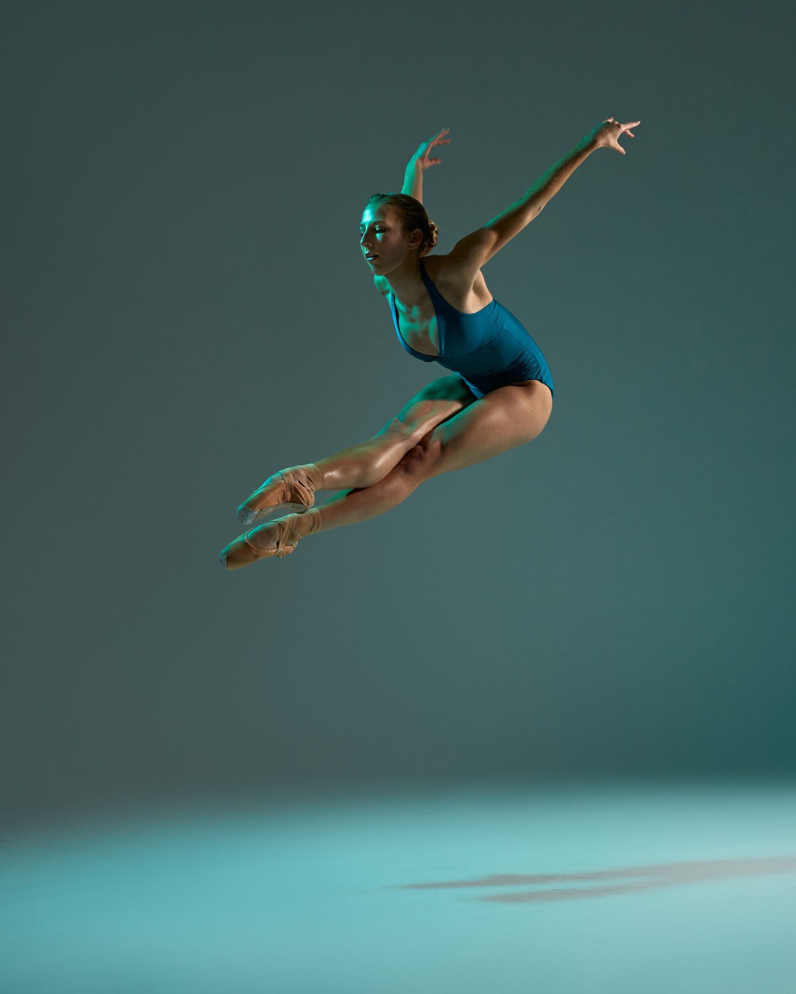 A female gymnast in a blue leotard jumps with arms and legs extended, against a plain background.