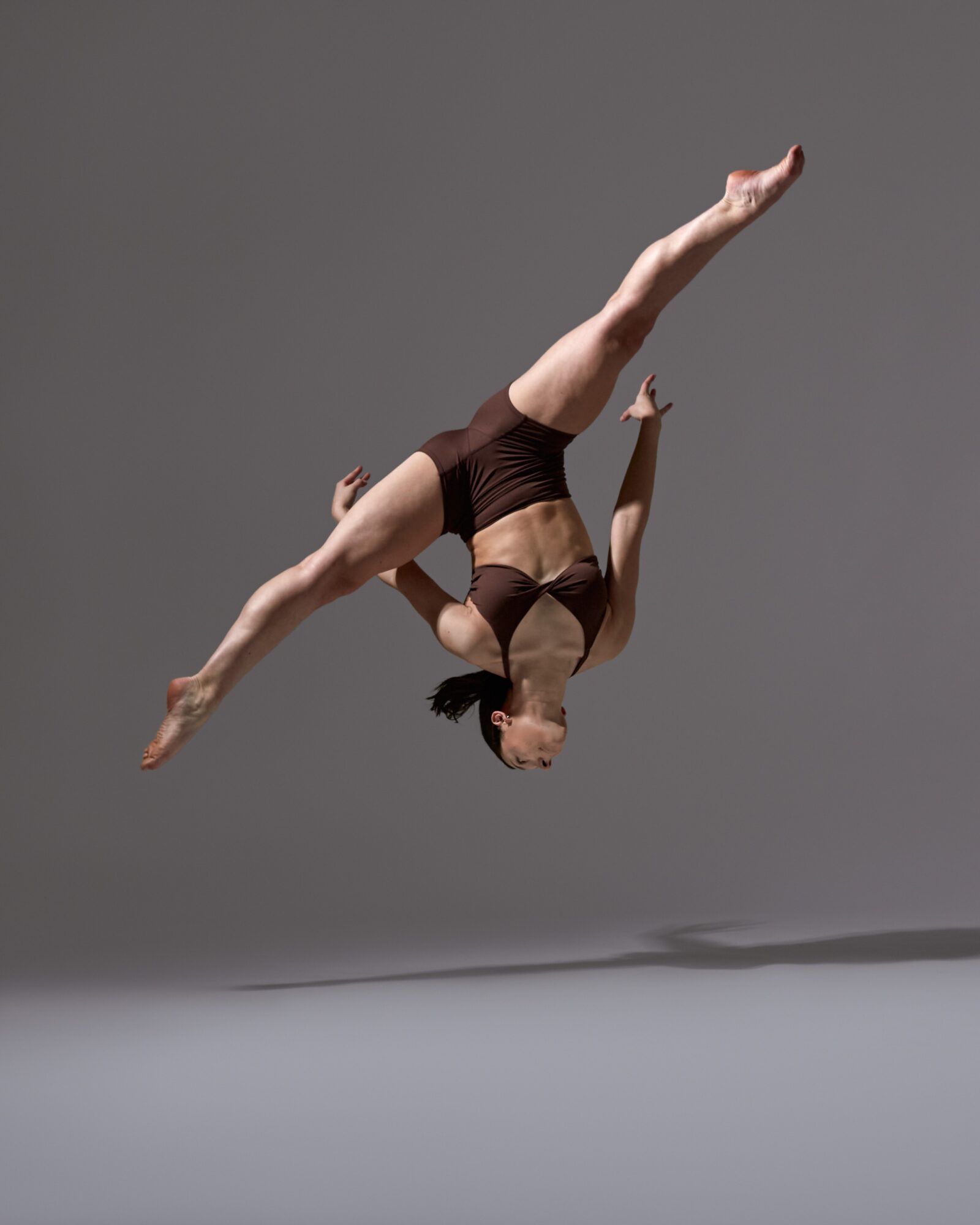 Female gymnast in mid-air, upside down with arms and legs extended, against a plain gray background.