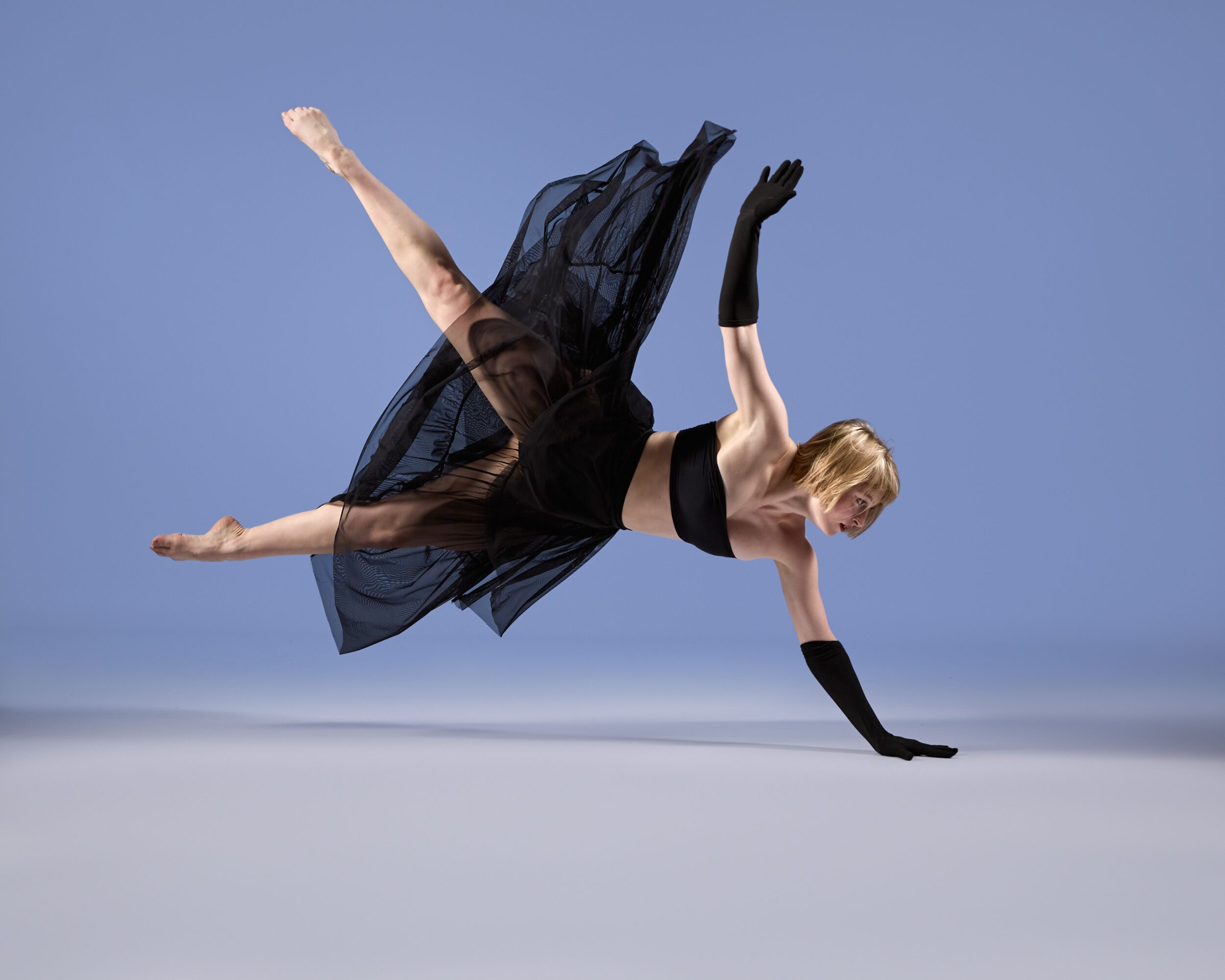 A woman in a black dress and gloves performs a dance move on a plain background.