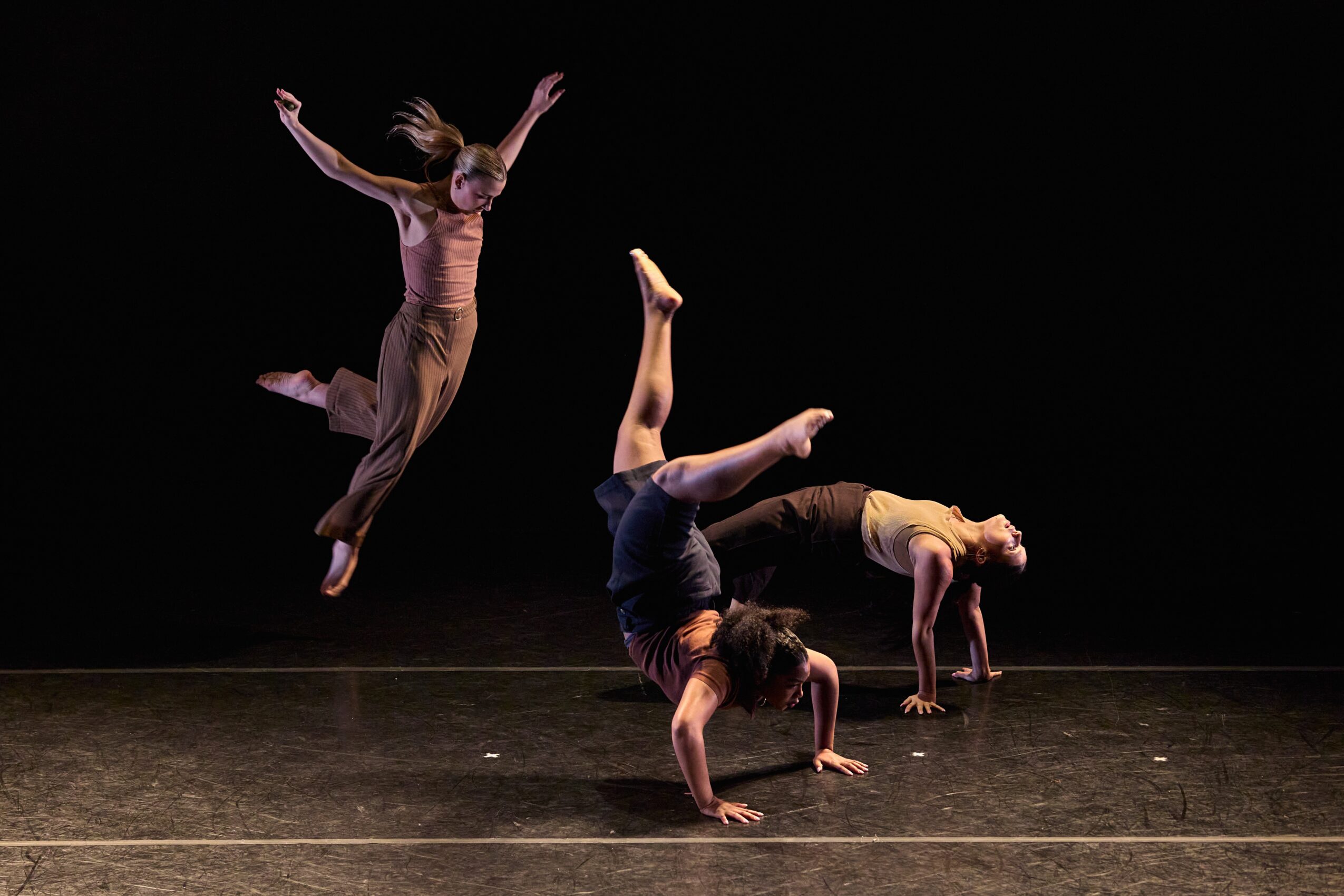 Three dancers perform on stage, one jumping, two in handstands, against a black background.