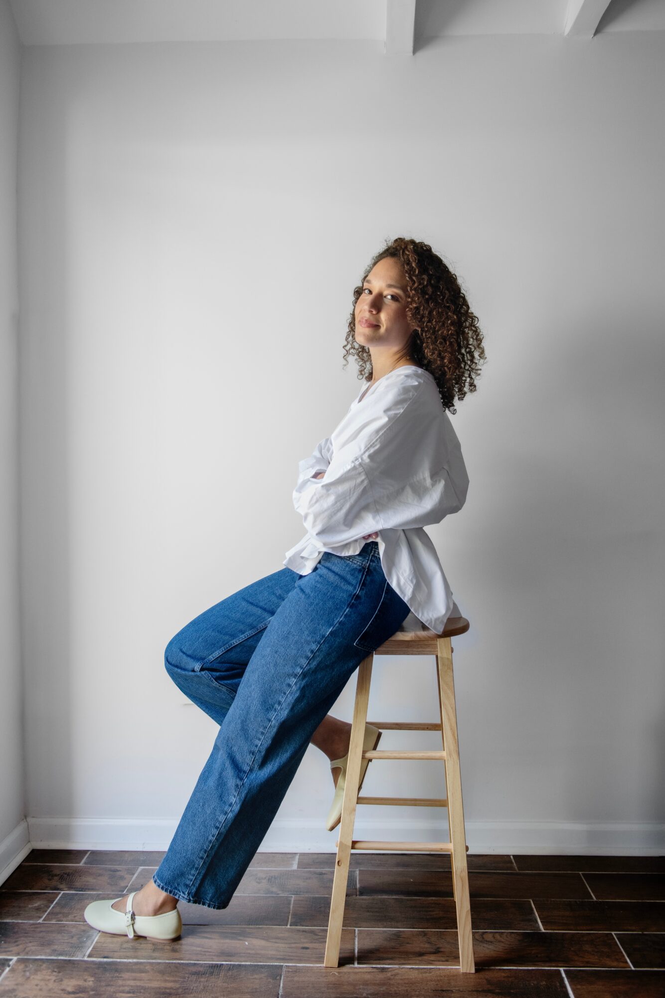 Woman with curly hair sitting on a wooden stool against a plain white wall, wearing a white shirt and blue jeans.