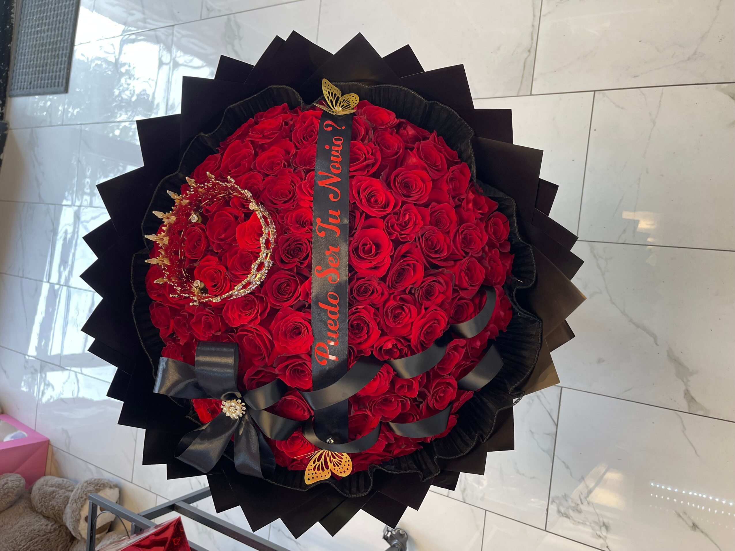 Round bouquet of red roses with black ribbon, gold butterfly, and decorative elements, wrapped in black paper, on a tiled floor.