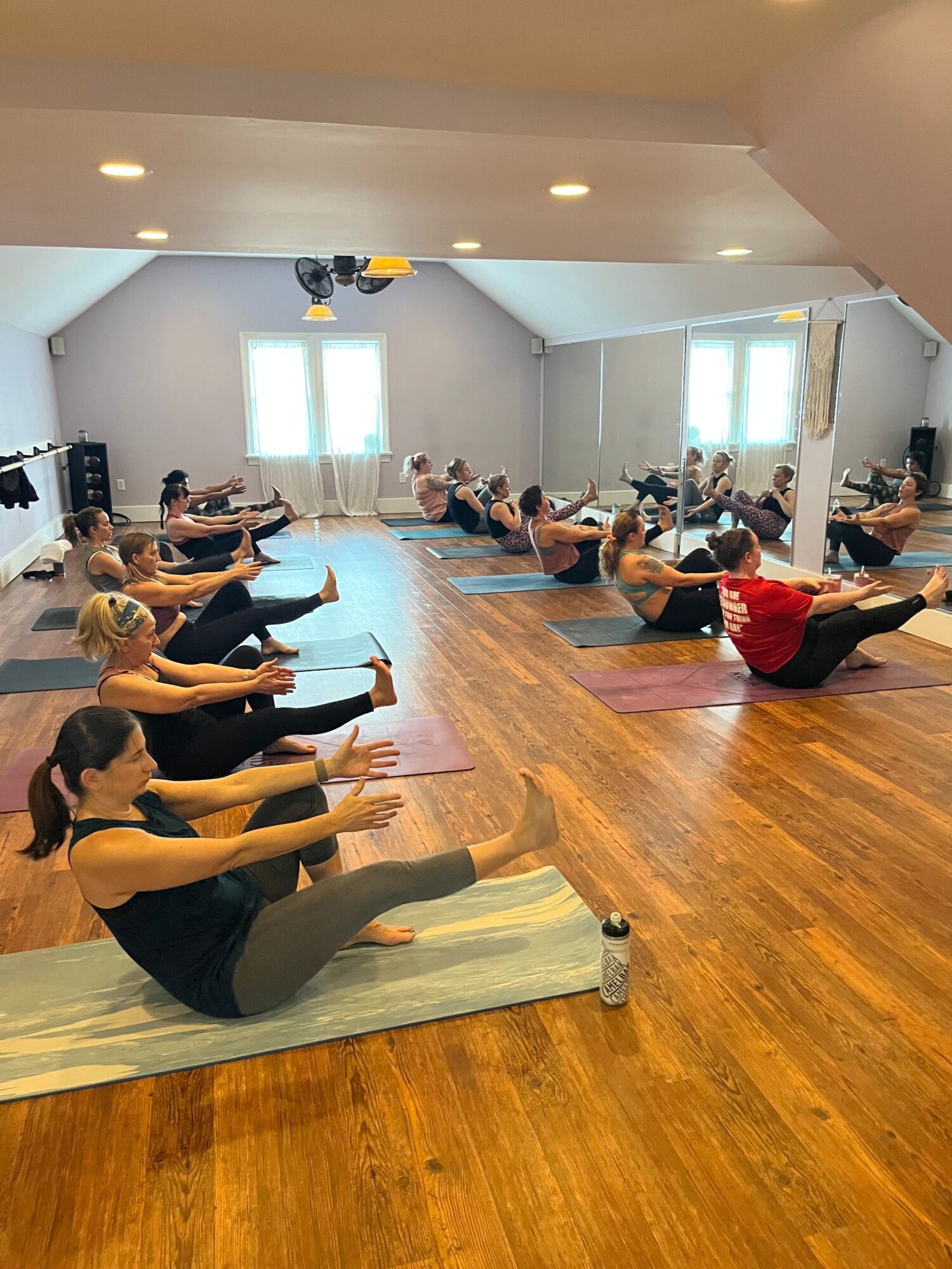 People participating in a yoga class in a spacious room with wooden floors and large windows, some sitting and others lying on mats.
