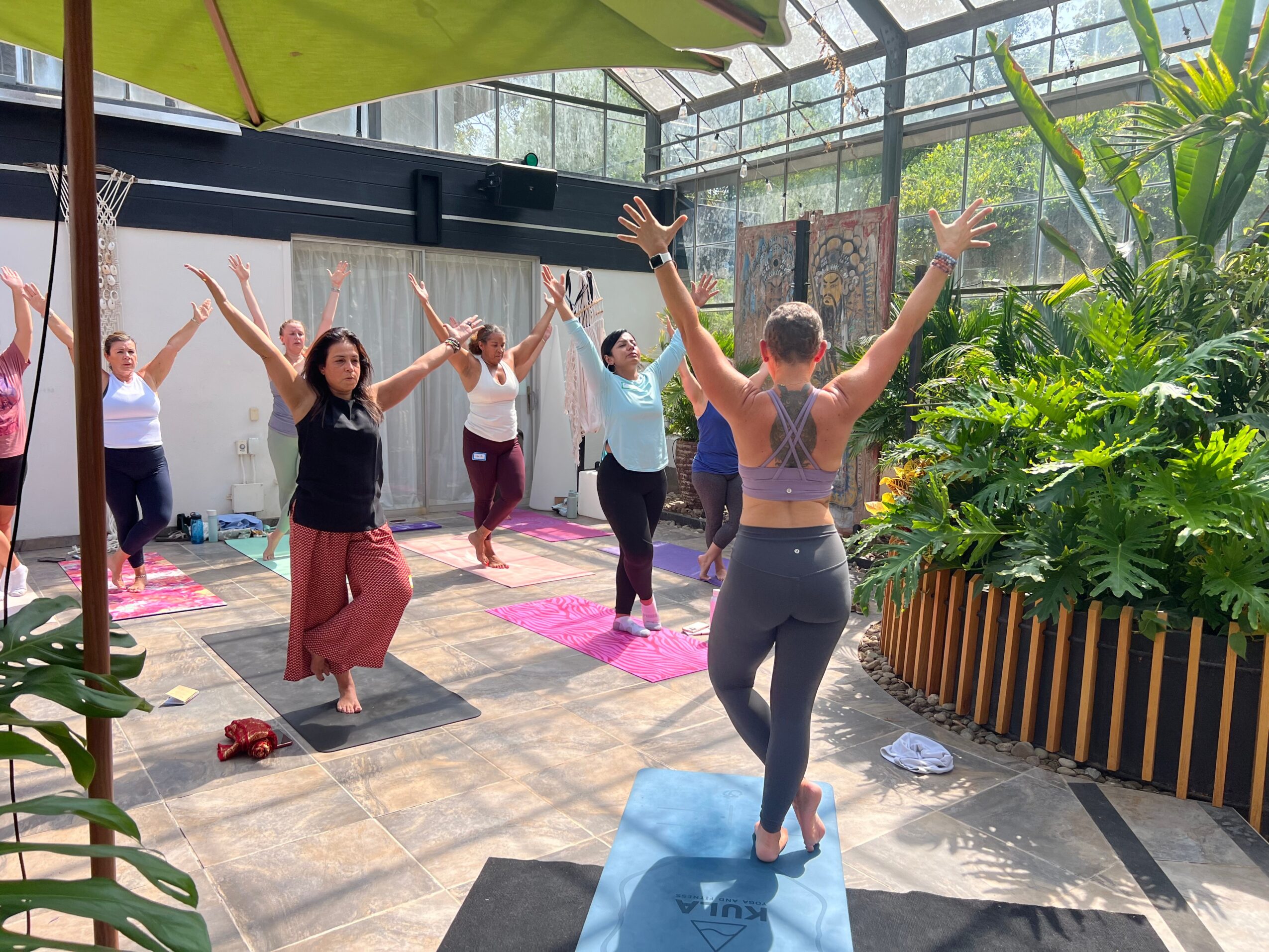 Group of people practicing yoga in a greenhouse with plants and sunlight, led by an instructor with arms raised, on colorful mats.