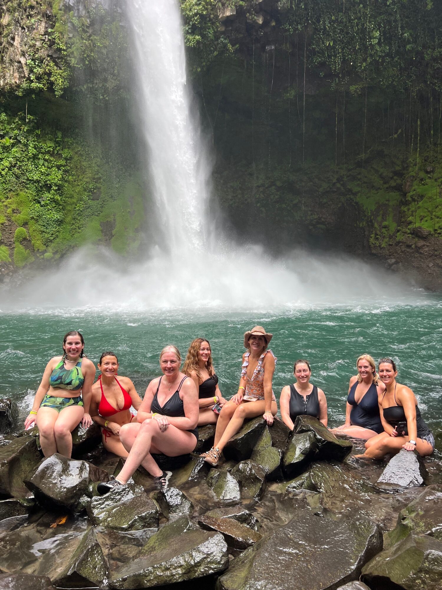 Group of people sitting on rocks by a waterfall in a lush green environment.