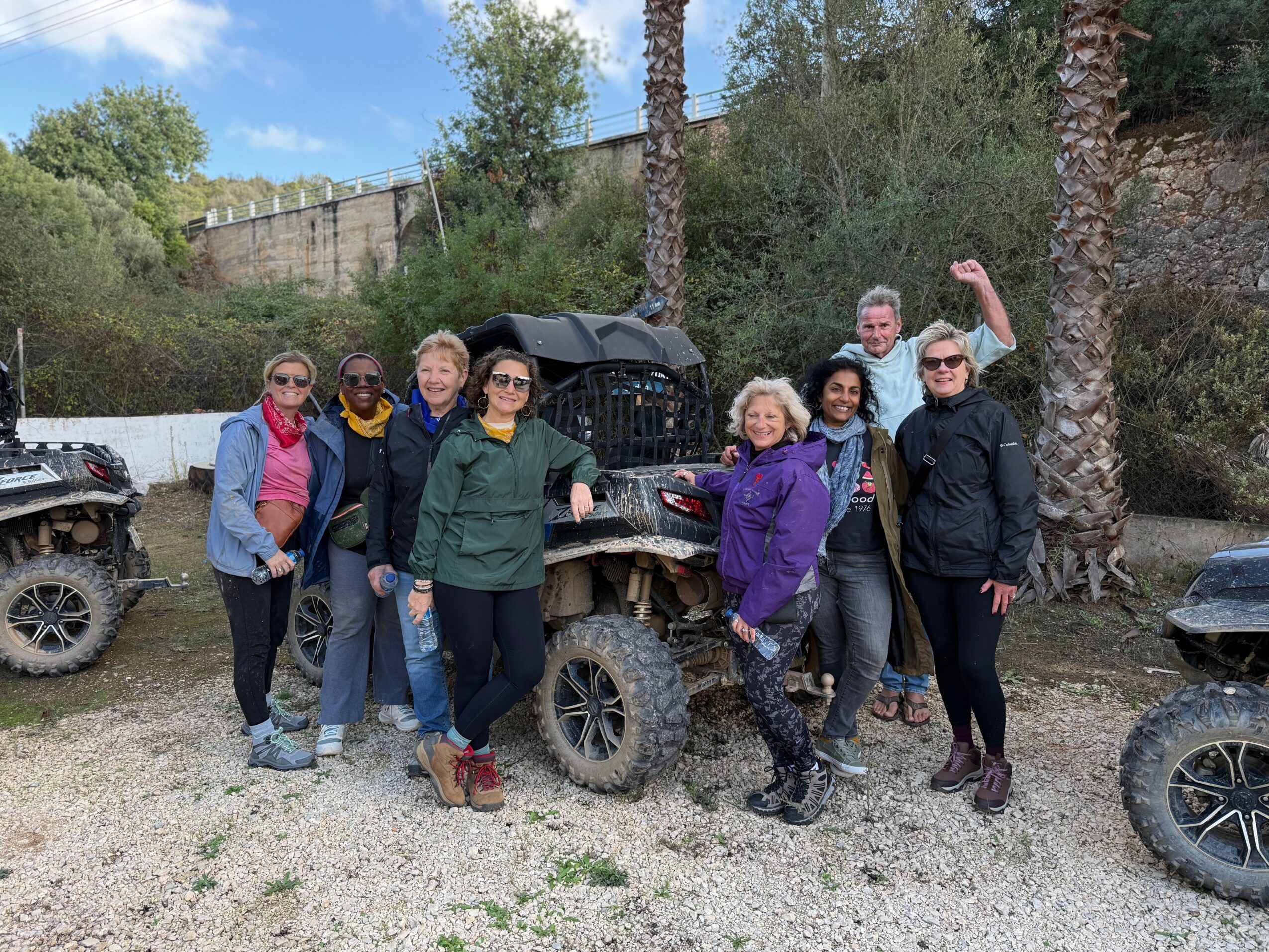 Group of eight people standing outdoors near trees and off-road vehicles, some holding umbrellas, smiling at the camera.
